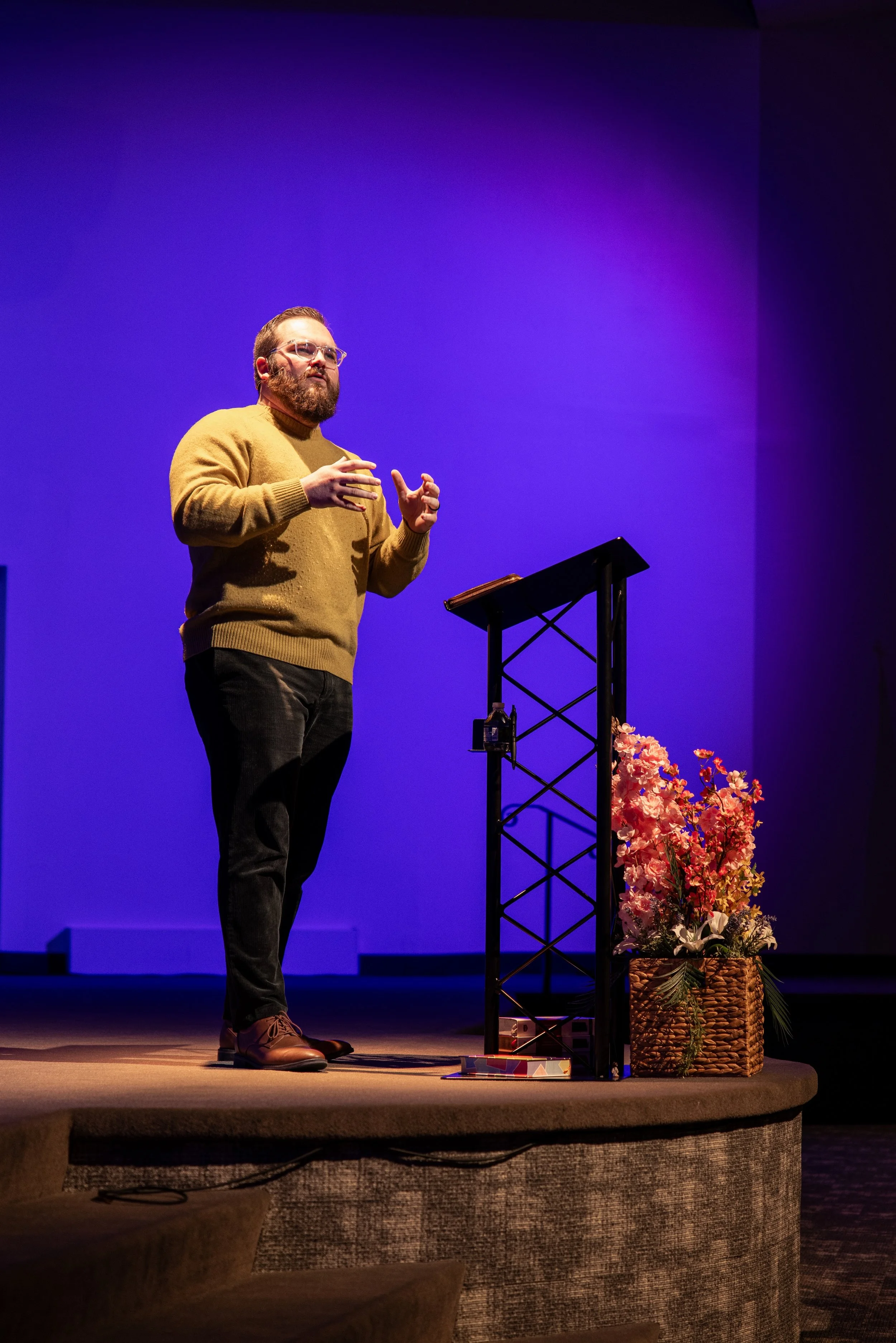 A man with glasses, beard, and wearing a brown sweater stands on a stage with purple lighting, speaking in front of a lectern beside a basket of flowers.