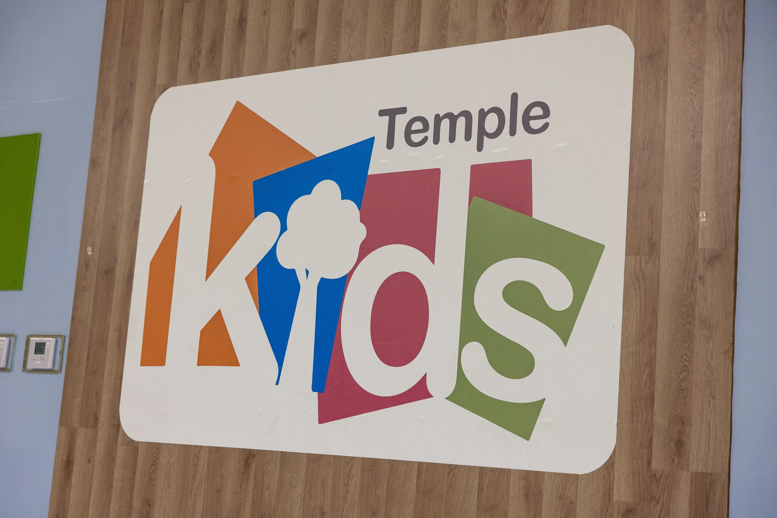 Sign with the text 'Temple Kids' in colorful letters and shapes, on a wooden wall.