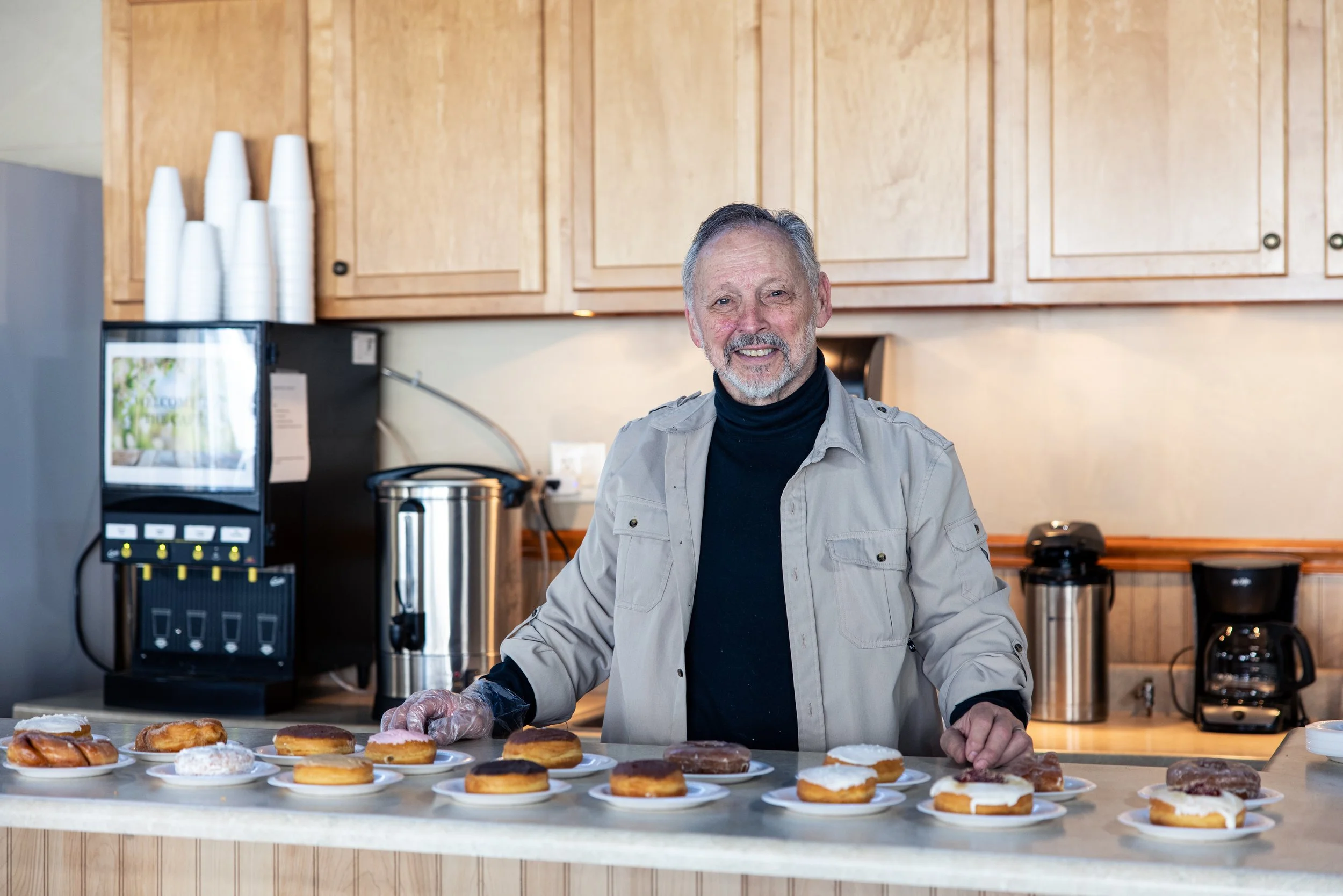 An older man with gray hair and a beard standing behind a table with various donuts on plates, smiling at the camera in a kitchen settings.