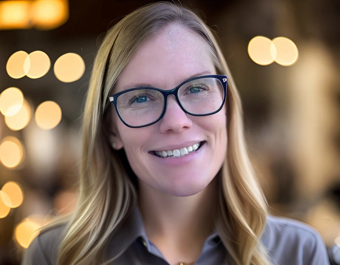 A smiling woman with long blonde hair and glasses, indoors with blurred warm lights in the background.
