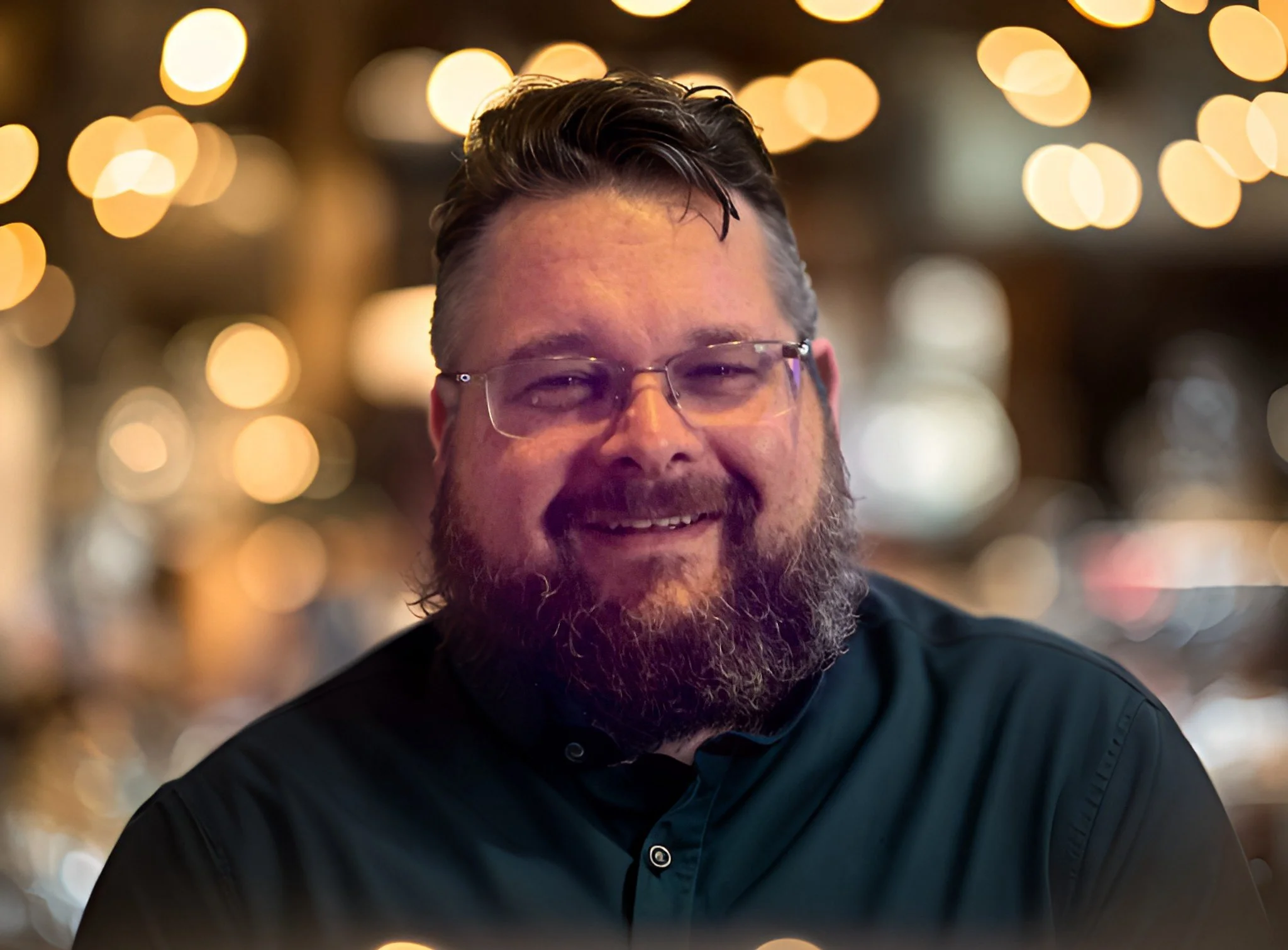 A smiling man with glasses and a beard, in a dimly lit setting with warm, circular bokeh lights in the background.