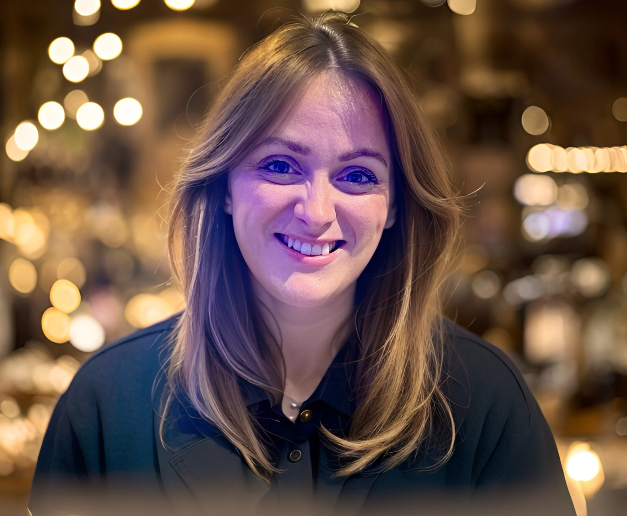A woman with long, light brown hair smiling at the camera in a warmly lit environment.