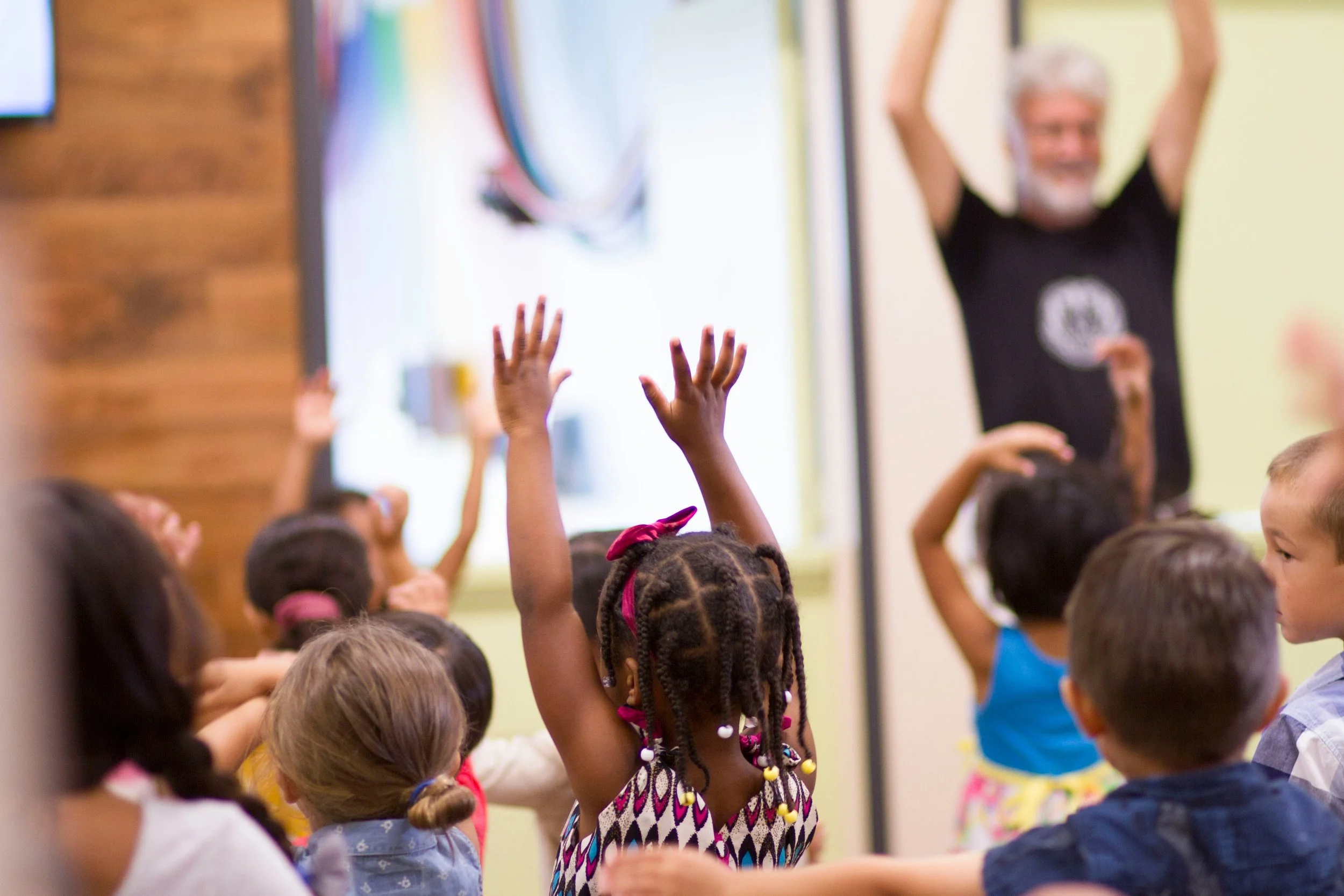 Children in a classroom with their hands raised, and a smiling instructor at the front.