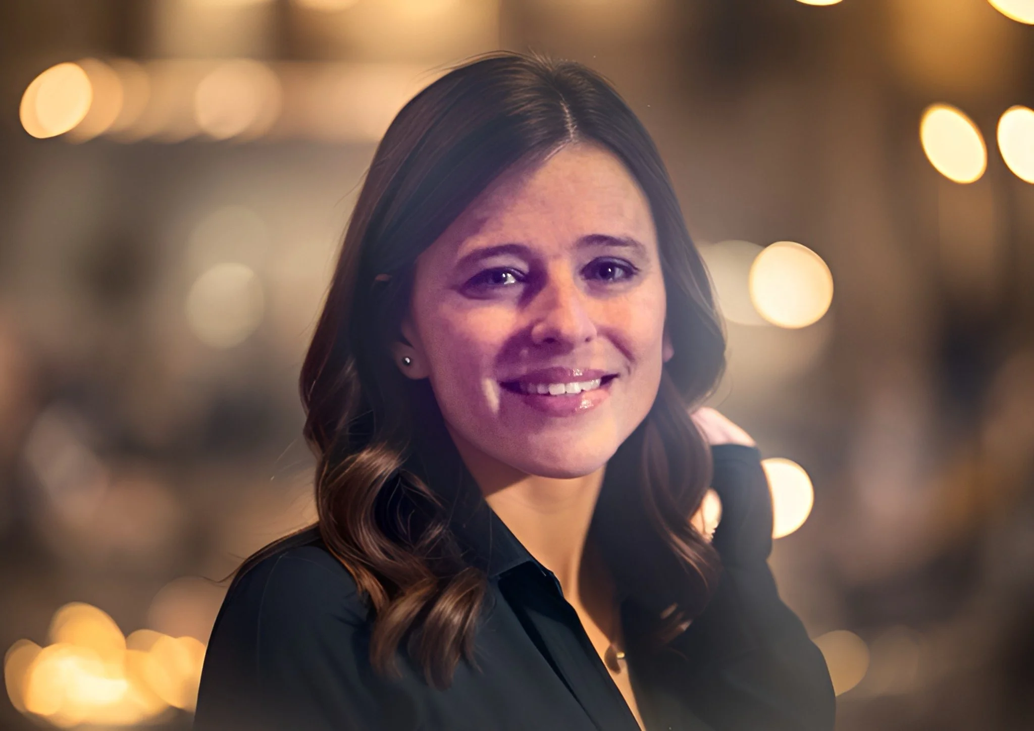 Portrait of a smiling woman with wavy brown hair and earrings, wearing a black top, with blurred bokeh lights in the background.
