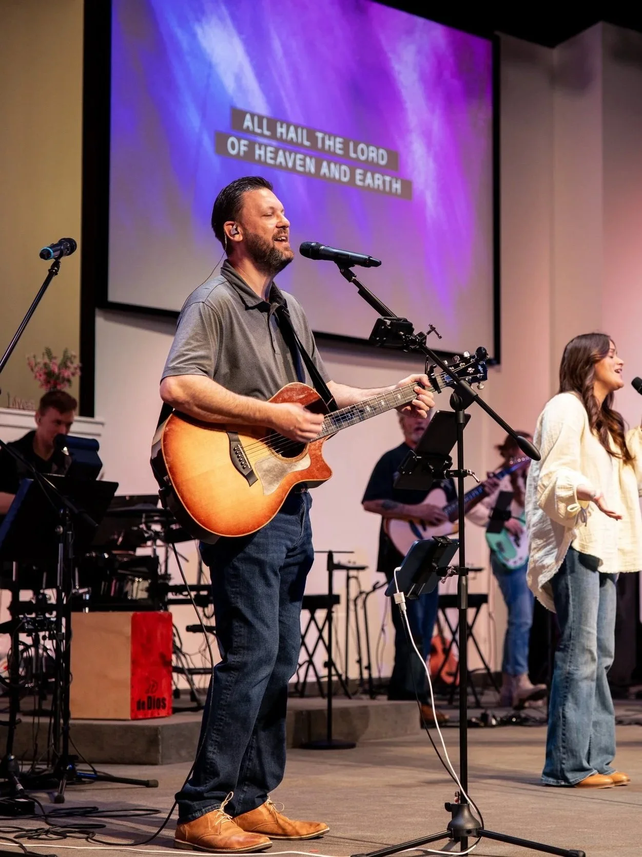 A man with a beard playing an acoustic guitar and singing on stage, with a band in the background. A woman is standing to the right singing. A large screen behind them displays the words 'All hail the Lord of heaven and earth'.