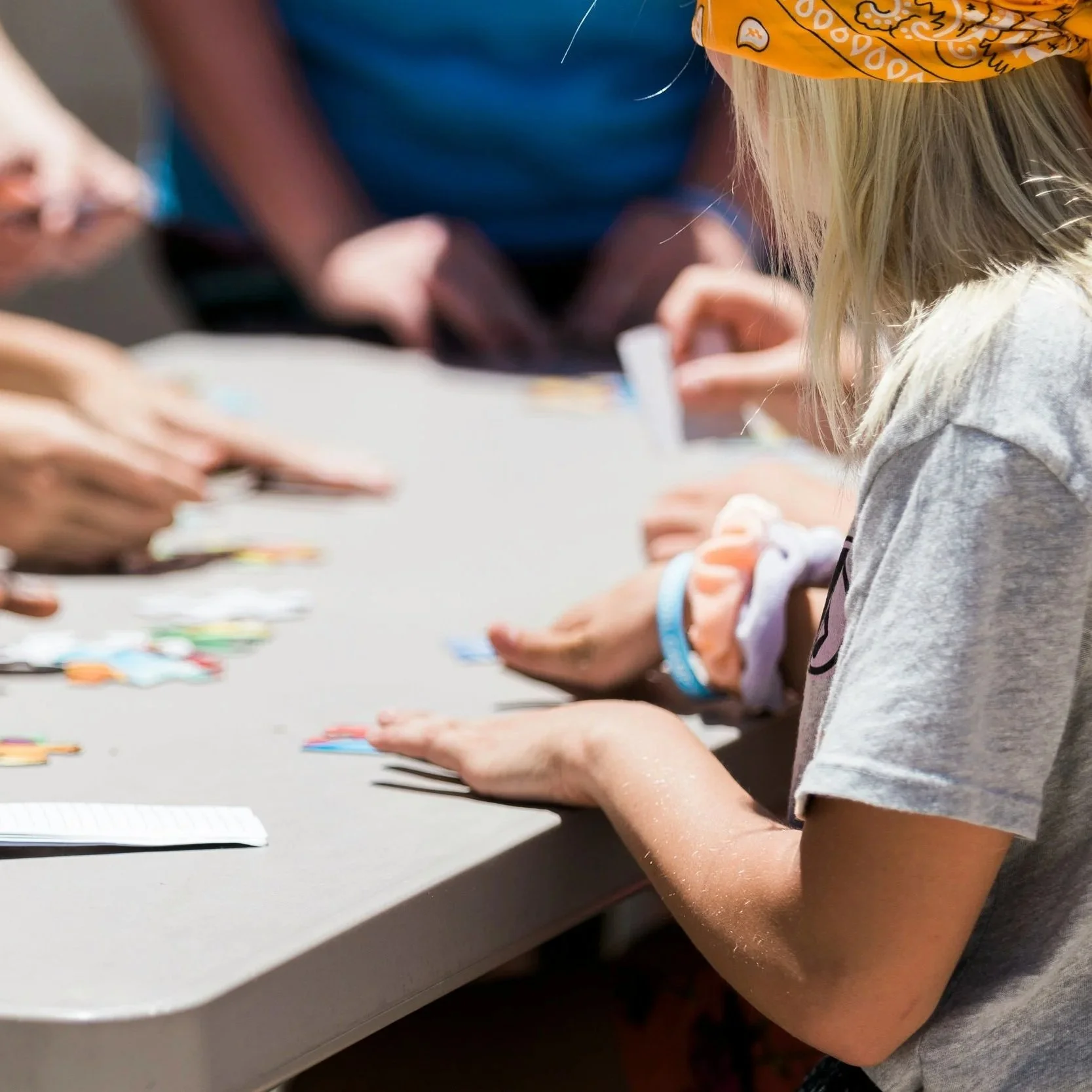 A young girl with blonde hair wearing a yellow bandana, gray t-shirt, and wristbands, playing a card game at a table during daytime.
