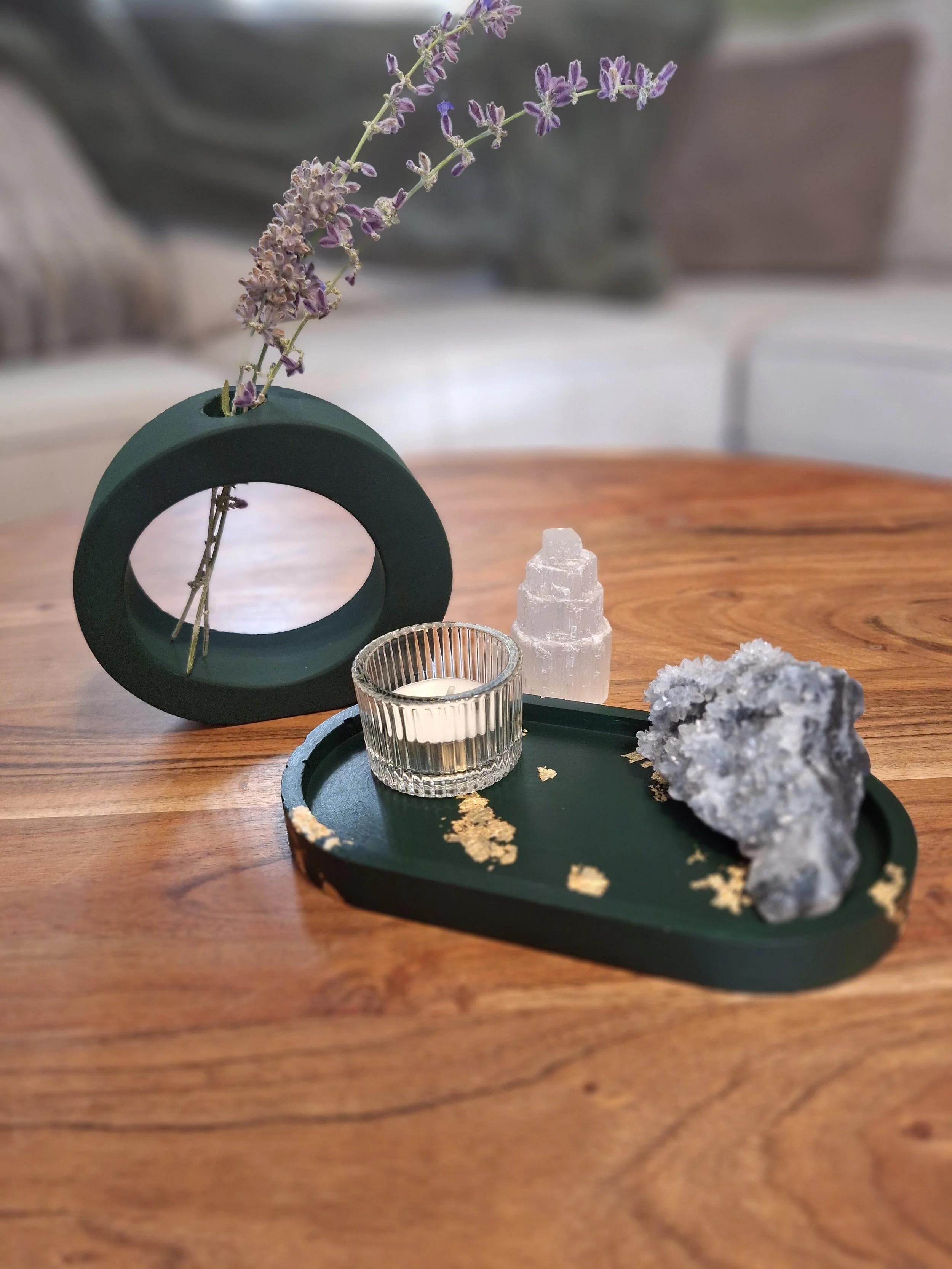 A wooden table with a decorative tray holding a candle in a glass holder, a crystal-like sculpture, and a large mineral or rock. Behind the tray, there's a dark green, circular, open-frame holder with a sprig of purple lavender flowers. In the background, out of focus, a gray sofa and some cushions are visible.