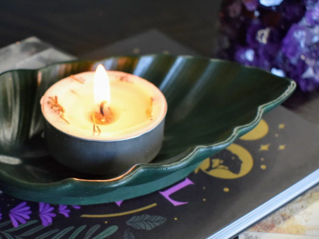 A lit candle in a black holder resting on a green leaf-shaped dish, placed on a decorative surface with a book and purple flowers nearby.
