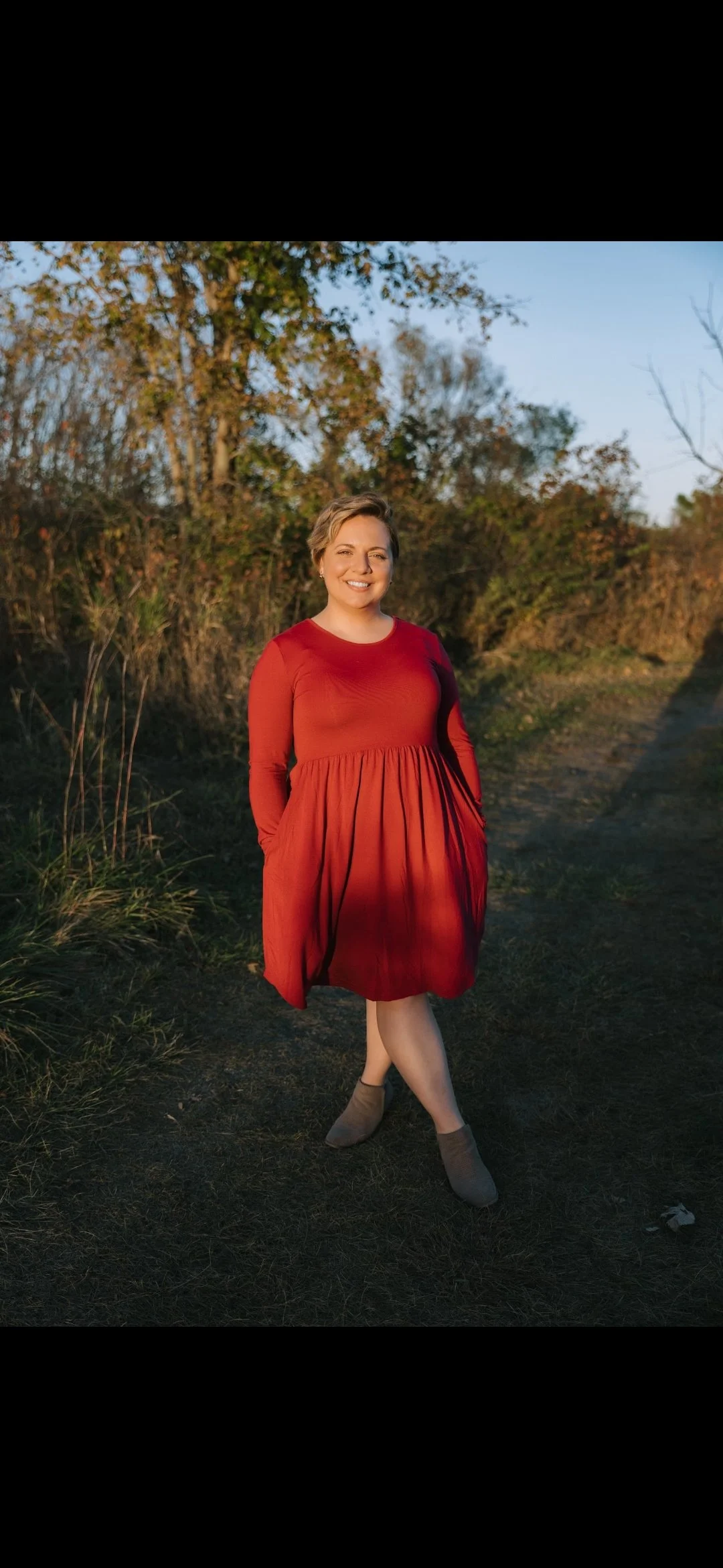 A woman standing outdoors on a dirt path in autumn, wearing a red dress and beige ankle boots, smiling at the camera with trees and foliage in the background.