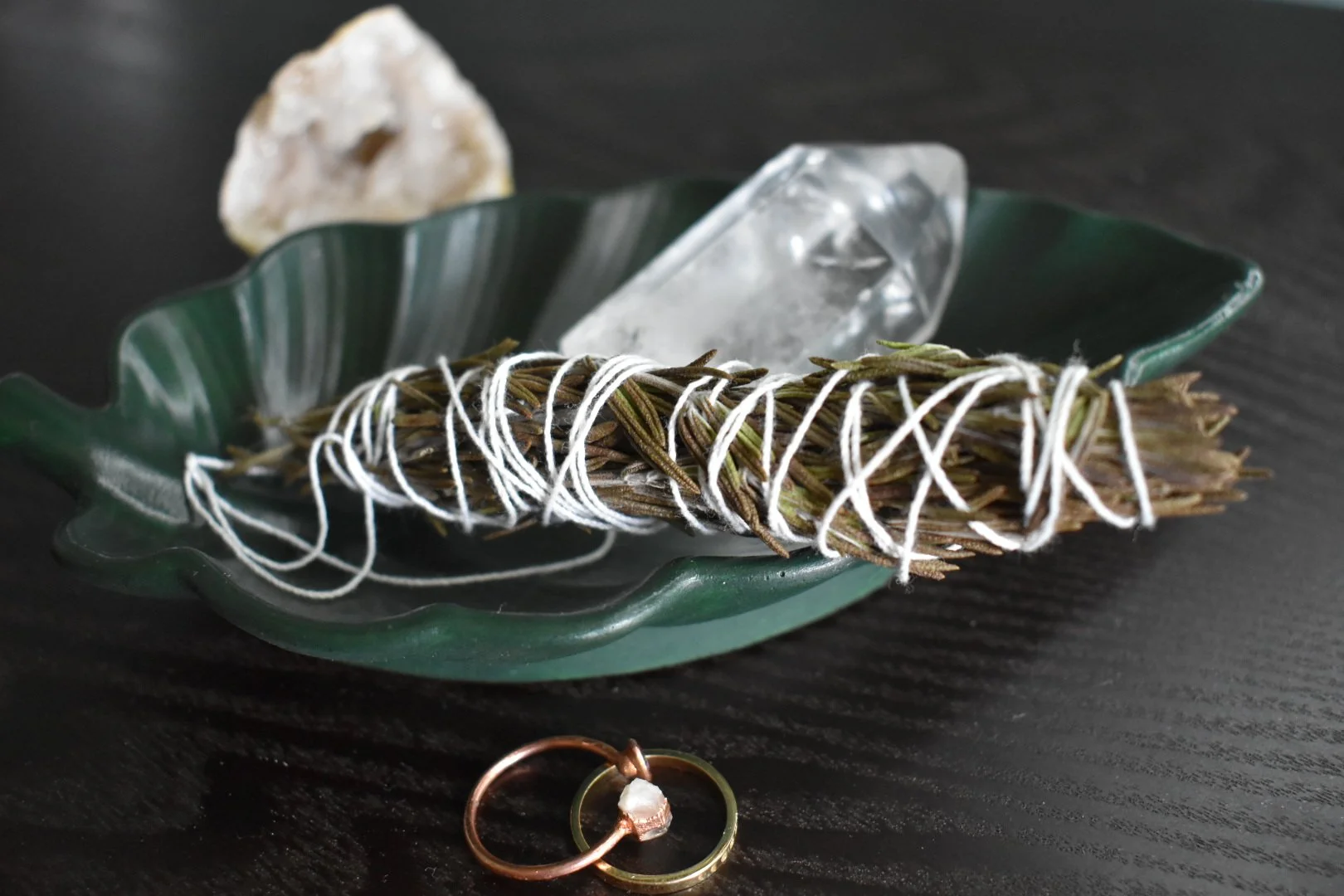 A thick bundle of sage wrapped with white string, resting on a green ceramic leaf-shaped dish. The dish contains a crystal and a shell fragment. Two rings are on the dark surface below the dish.