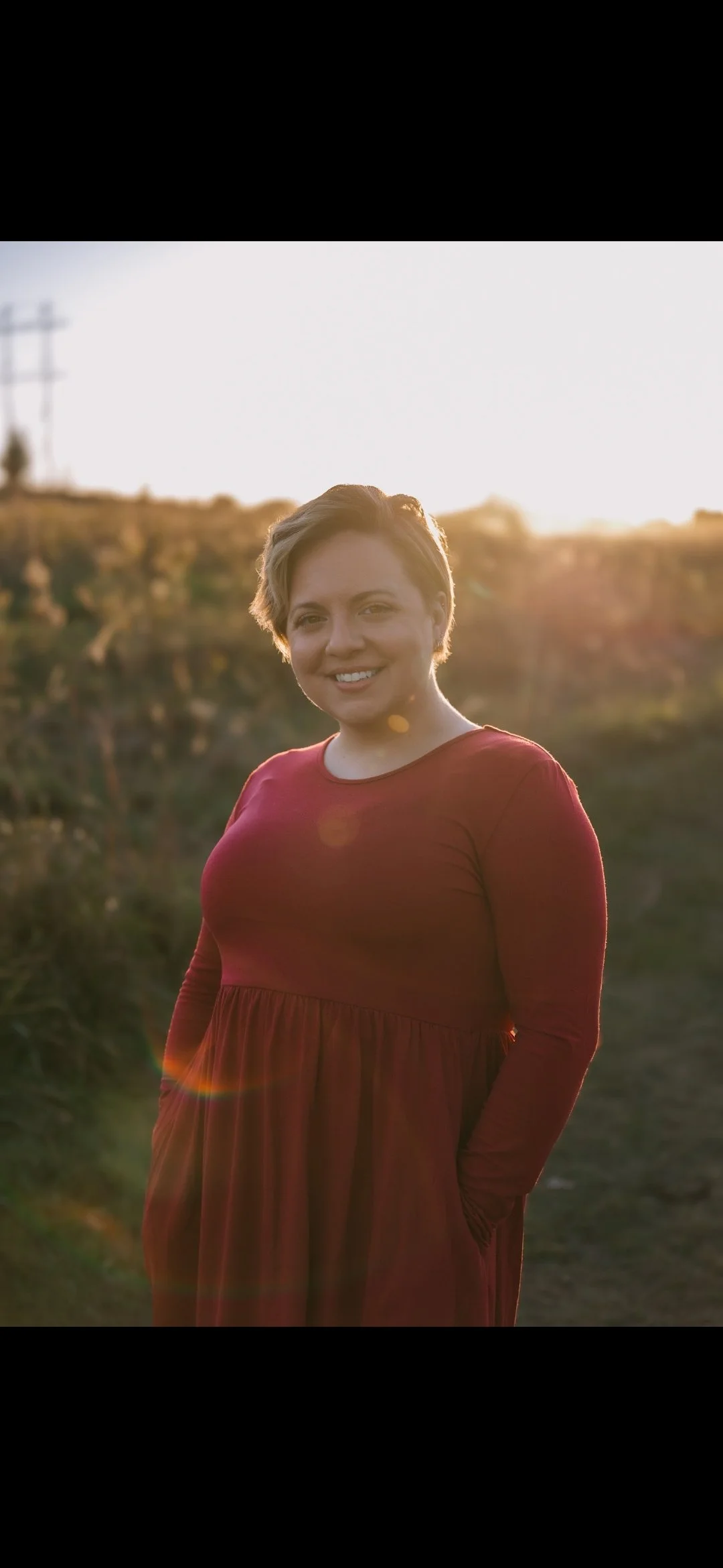 A woman with short brown hair wearing a long-sleeved red dress, smiling outdoors during sunset with a blurred landscape background and lens flare.