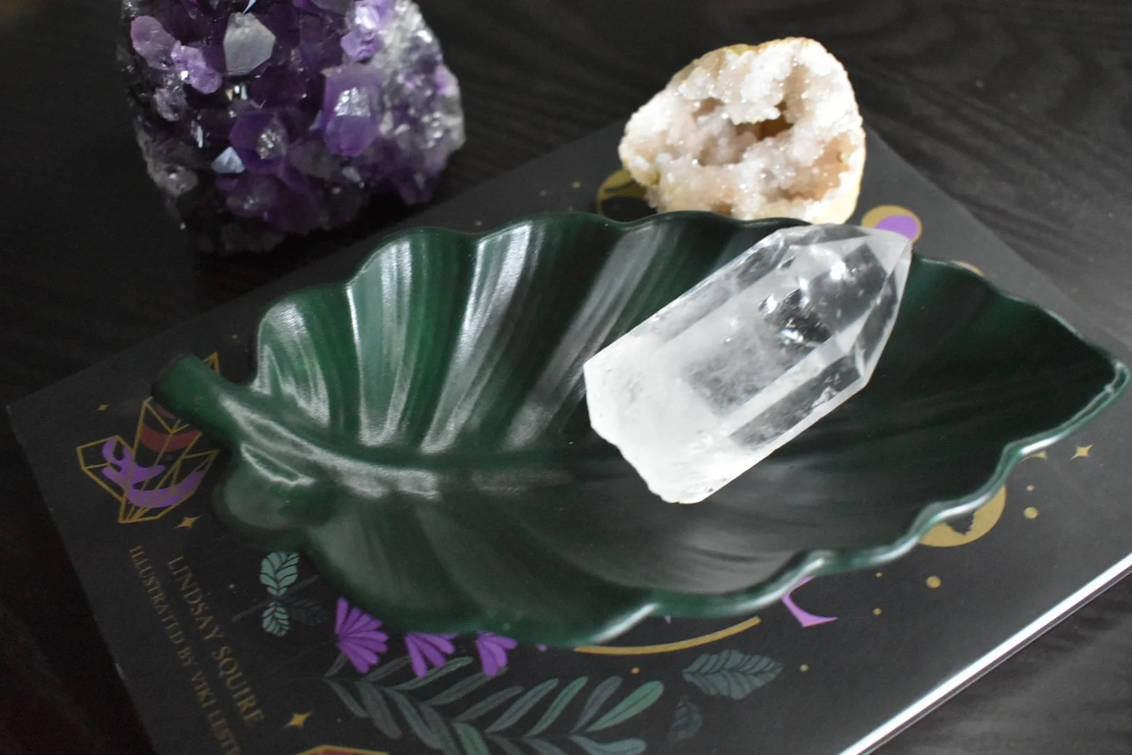 A clear quartz crystal, a purple amethyst mineral, and a white geode with crystals inside are on a green leaf-shaped dish.