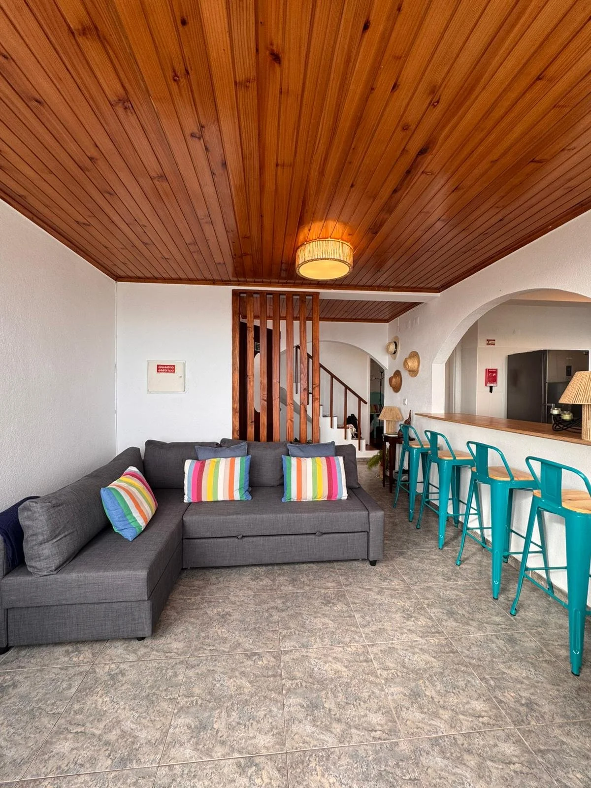 Living room with gray sectional sofa with rainbow-striped pillows, wooden ceiling, bar counter with turquoise chairs, and staircase in the background.
