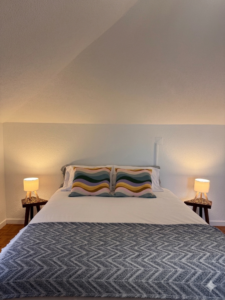 A neatly made bed with two pillows featuring colorful, wavy, striped pillowcases. On each side of the bed, there are small wooden nightstands with matching table lamps that have white shades. The bed has a textured, patterned gray blanket at the foot, and the background consists of a plain light-colored wall with a slight slant in the ceiling.