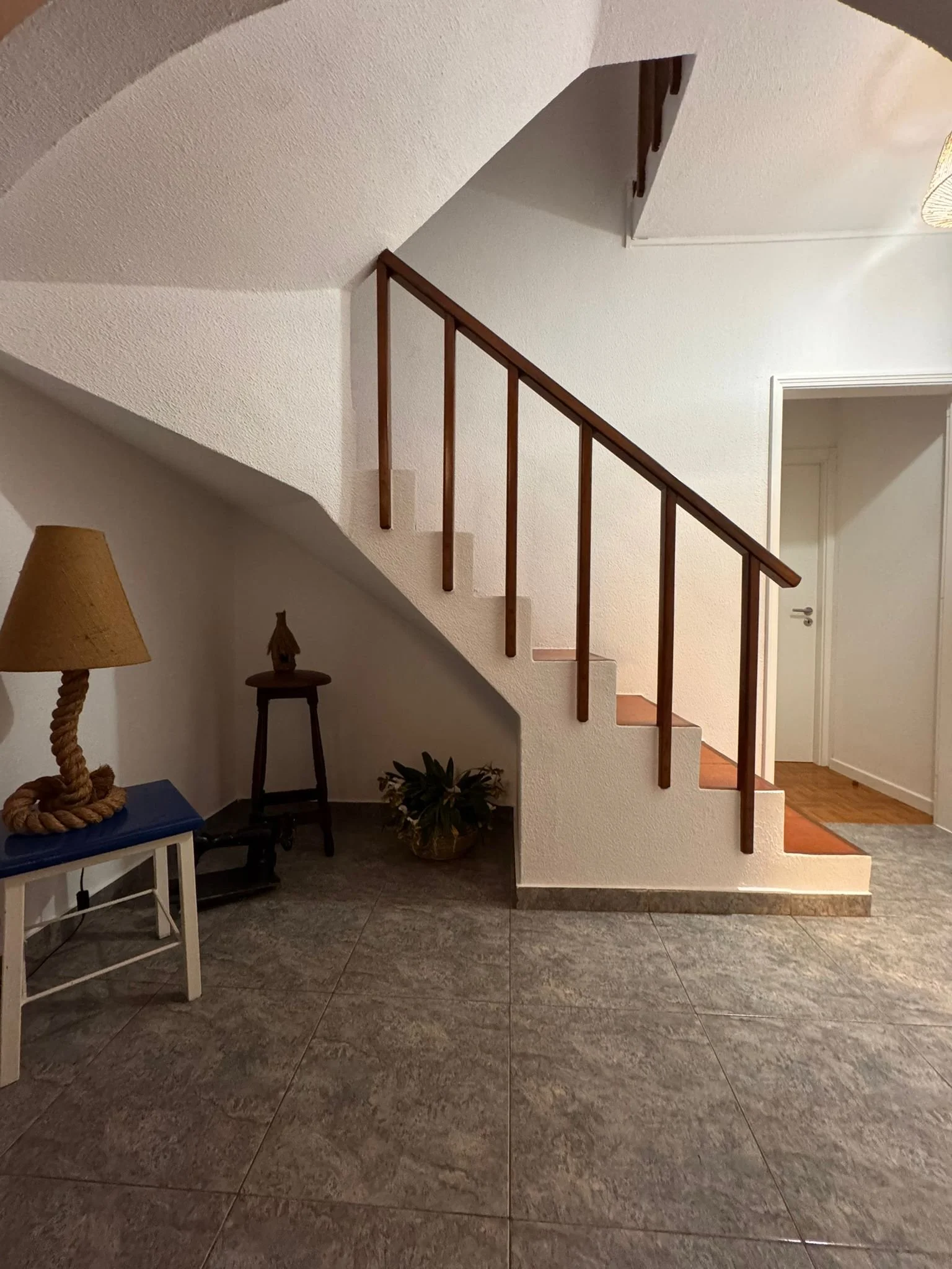 Interior view of a house entryway with a staircase featuring wooden handrails and beige steps, a small corner with a plant, a side table with a lamp, and a door leading to another room.