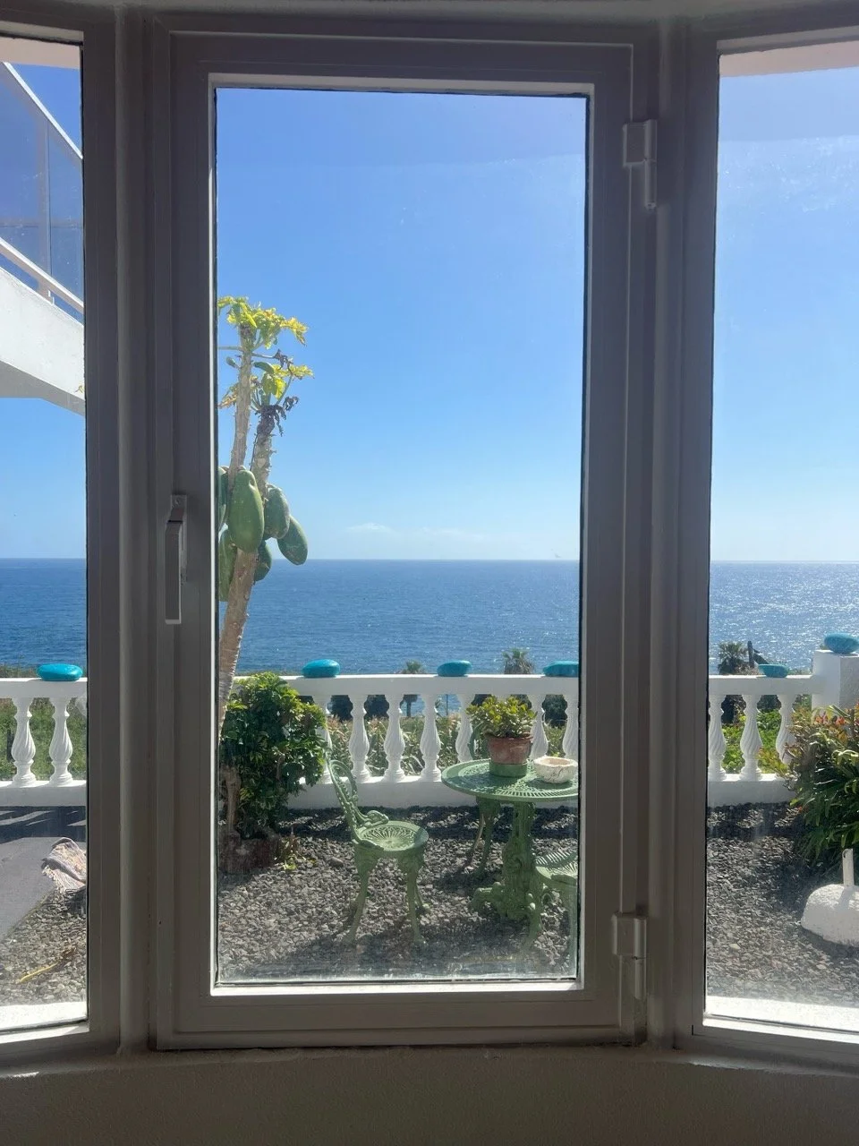 View of a sunny balcony with a potted cactus, green garden furniture, and the ocean in the distance, seen through a glass door.