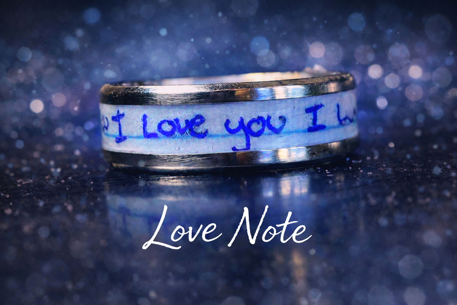 Close-up of a silver ring with a handwritten blue note wrapped around it that says "I Love You" against a sparkly dark background, with the words "Love Note" written below.