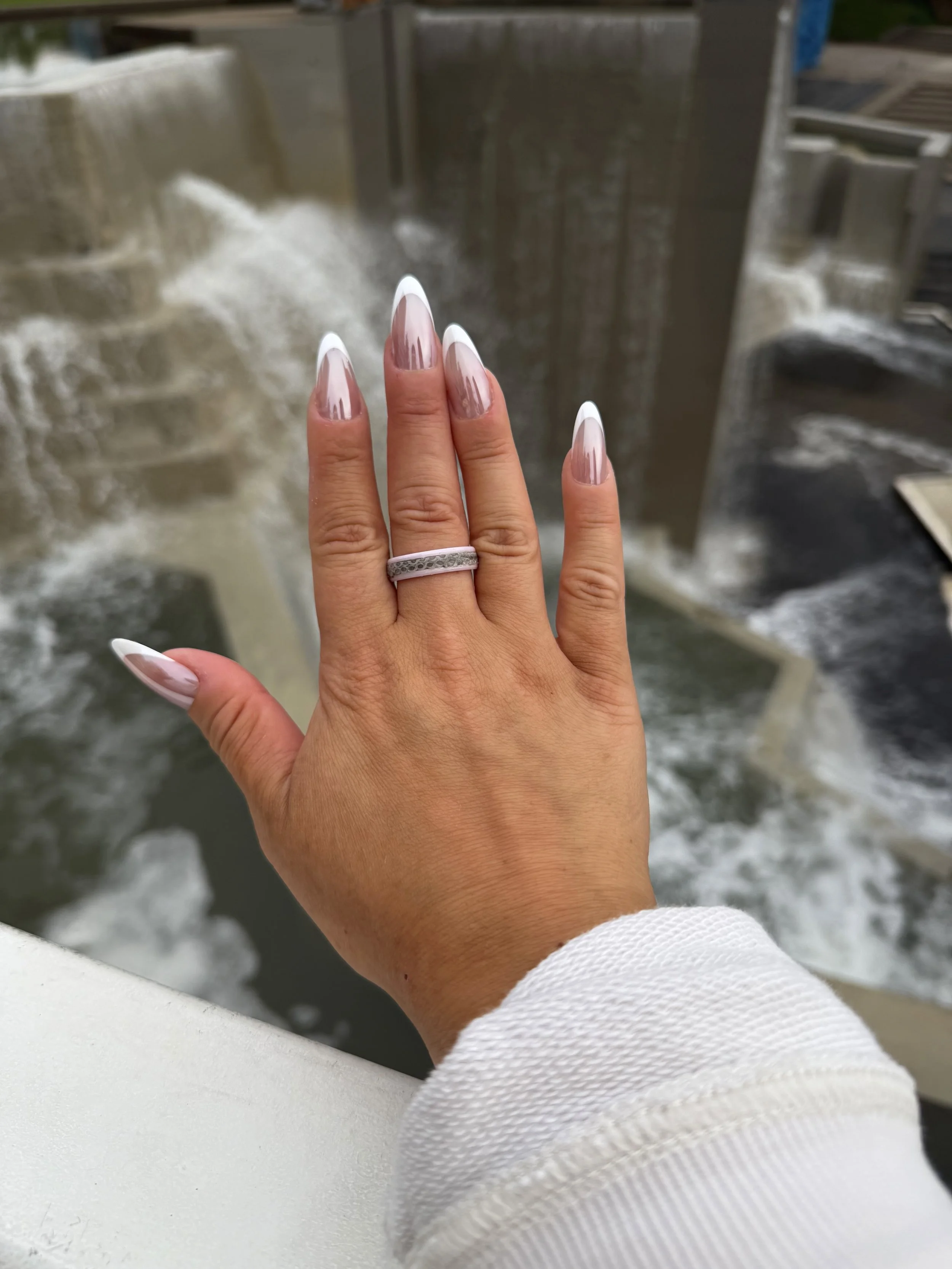 Close-up of a hand with manicured nails and a silver ring, reaching toward a water feature in an outdoor setting.