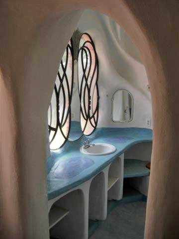 A whimsical bathroom with a curved architectural design, featuring a blue countertop with a built-in sink, decorative stained glass windows with swirling patterns, and an arched mirror on the wall.