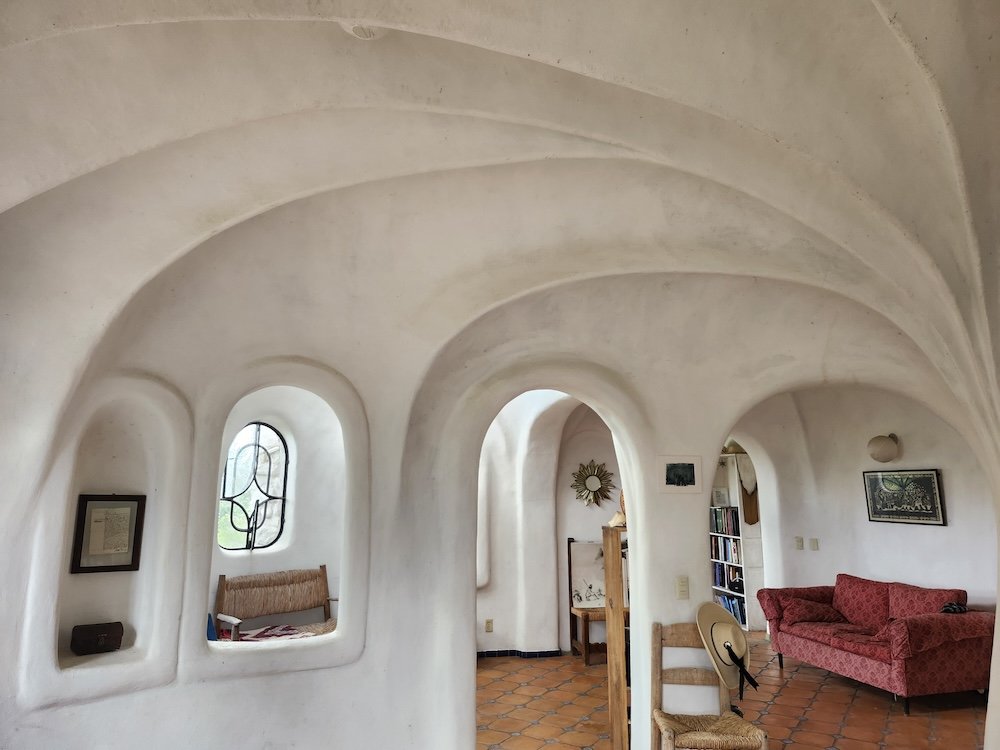 Interior of a house with white, curved plaster walls and ceiling, terracotta tile floor, small arched windows with decorative ironwork, a pink patterned sofa, wooden chairs, and framed artwork.