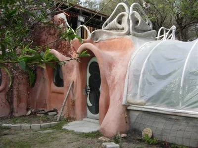 A whimsical pink and beige house with an organic, curvy design, situated outdoors among trees and greenery.
