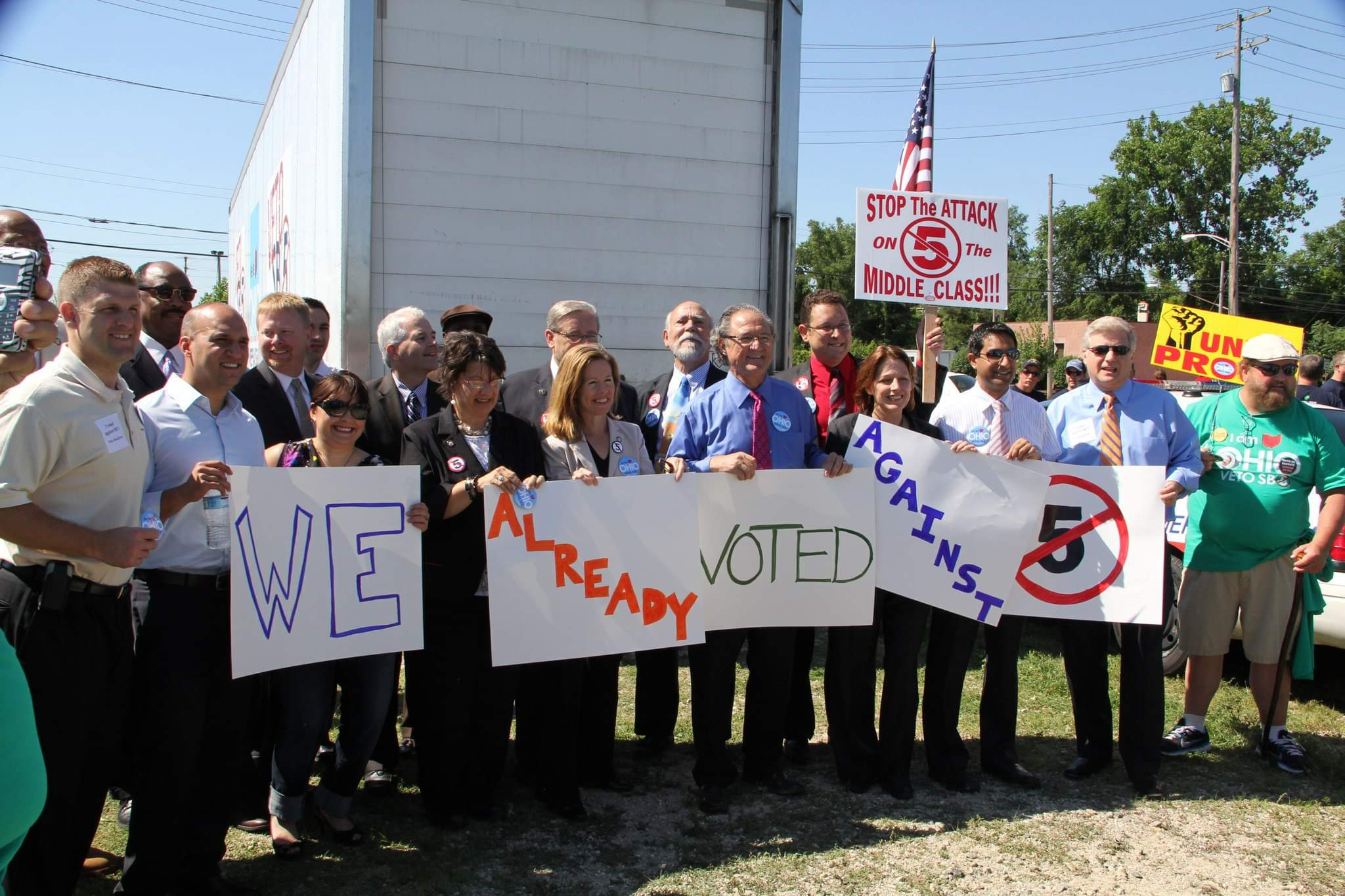 Group of people protesting with signs against the attack on the middle class, including signs that say "We are ready," "Voted against," and stop the attack on the middle class. One person holds a sign with a crossed-out 5 inside a circle, and there are people in the background with signs and flags.