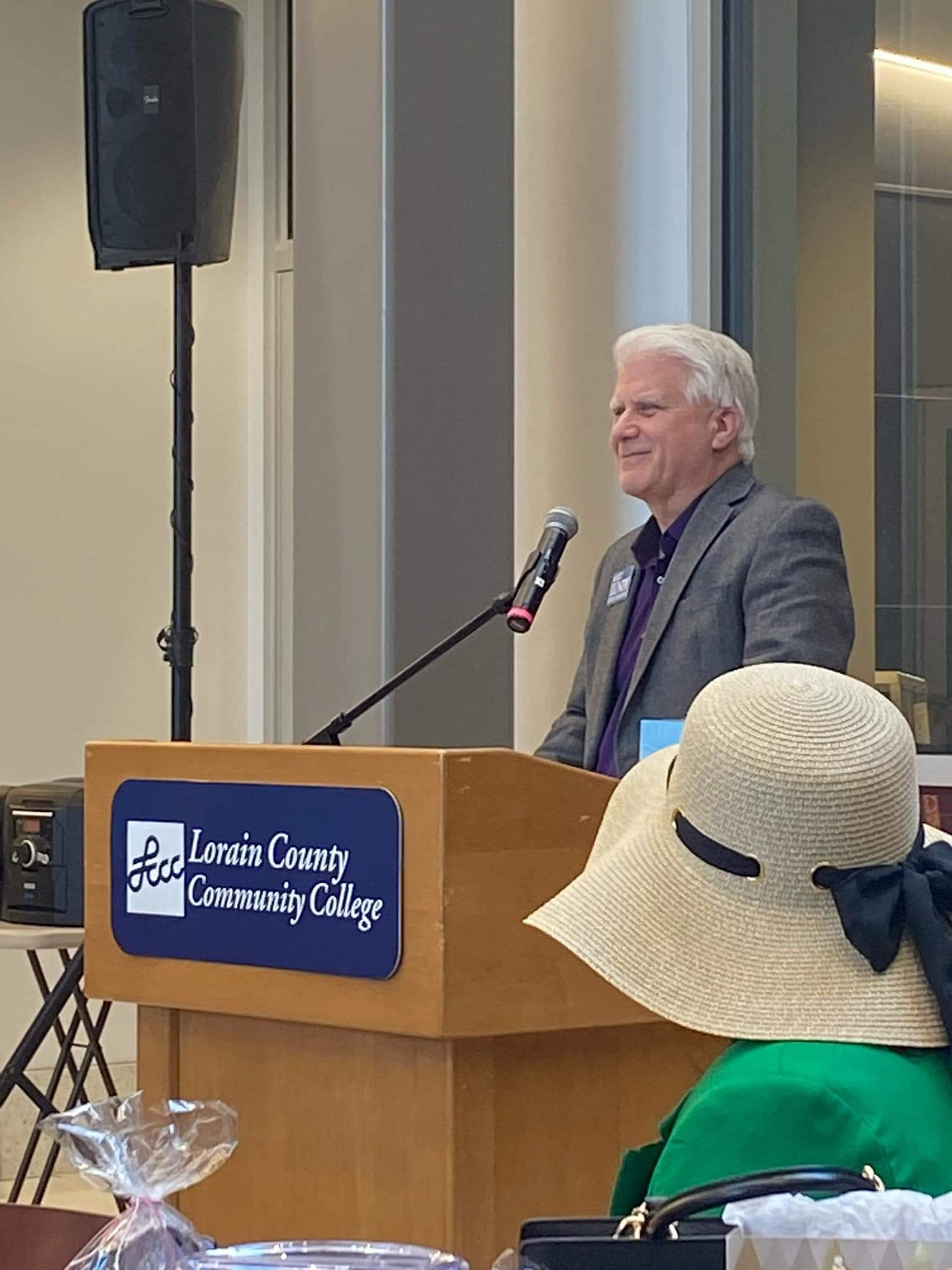 Matt Lundy is giving a speech at a podium with a Lorain County Community College sign. He is wearing a gray blazer and speaking into a microphone to an audience.