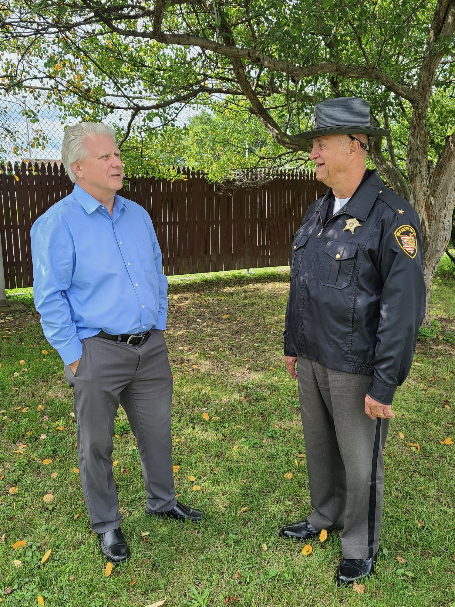Matt Lundy and a Lorain County Sheriff's Deputy are standing and chatting under a tree with sparse leaves, a grassy yard, and a wooden fence in the background.