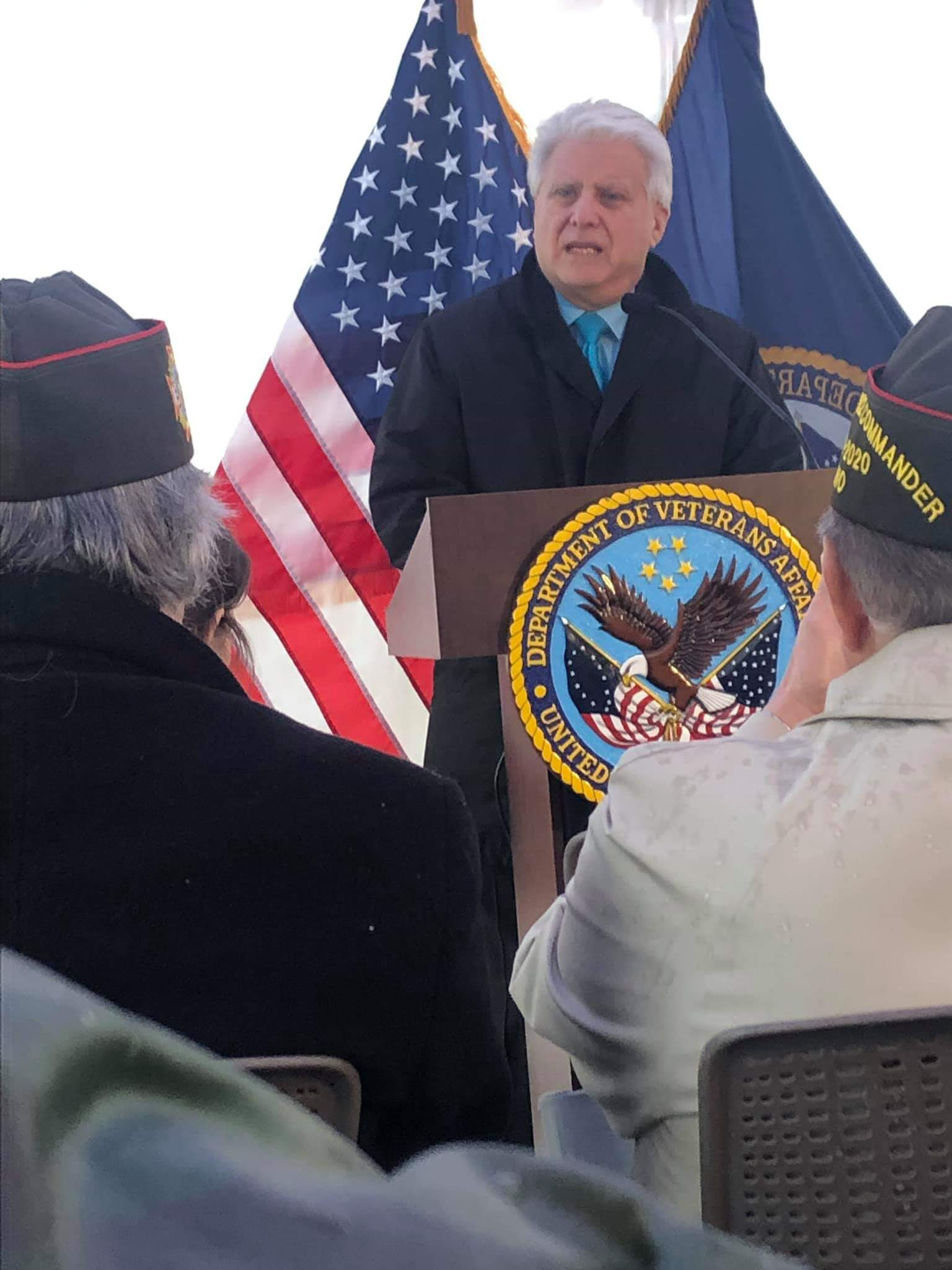 Matt Lundy stands at a podium with the seal of the Department of Veterans Affairs. Behind him is the American flag and the Dept. of Veterans Affairs flag. He is speaking to Veterans whose backs are to the camera.