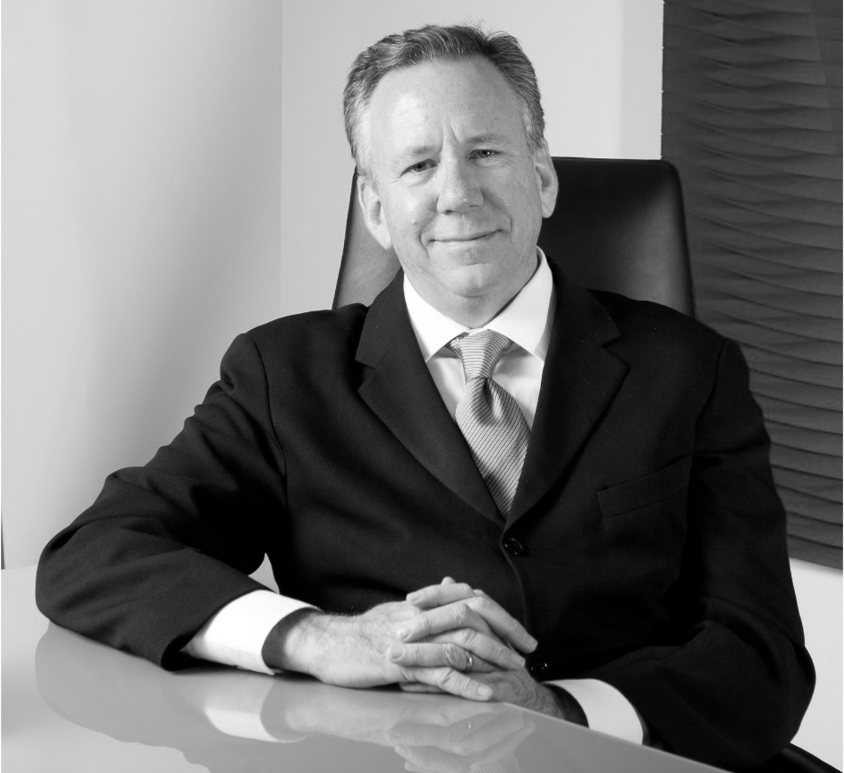 Black and white photo of a middle-aged man in a suit, sitting at a table with his hands clasped, smiling slightly, in an office setting.