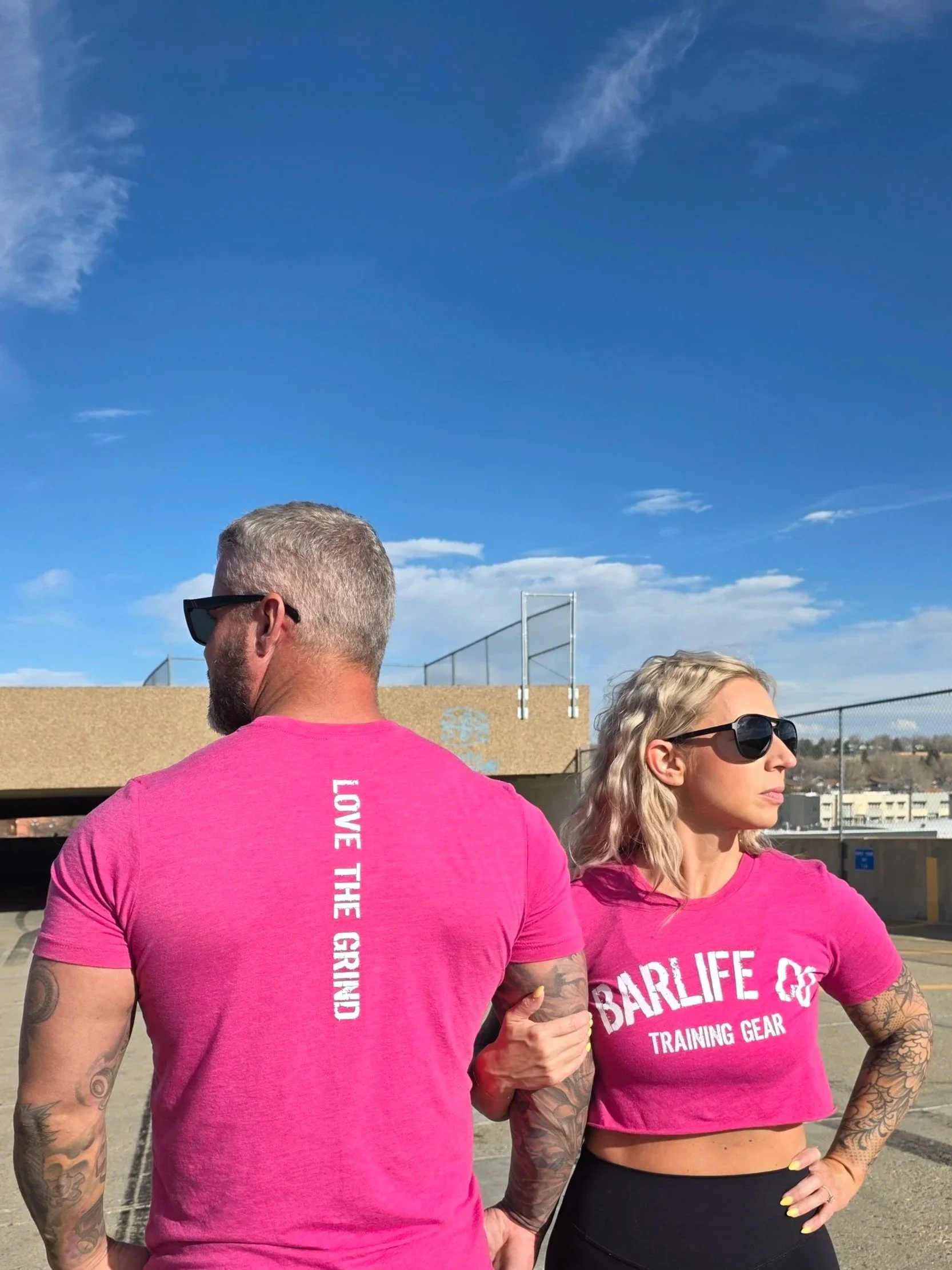 A man and woman standing on a rooftop parking lot, wearing pink workout shirts, looking off to the side with sunglasses, under a partly cloudy sky.
