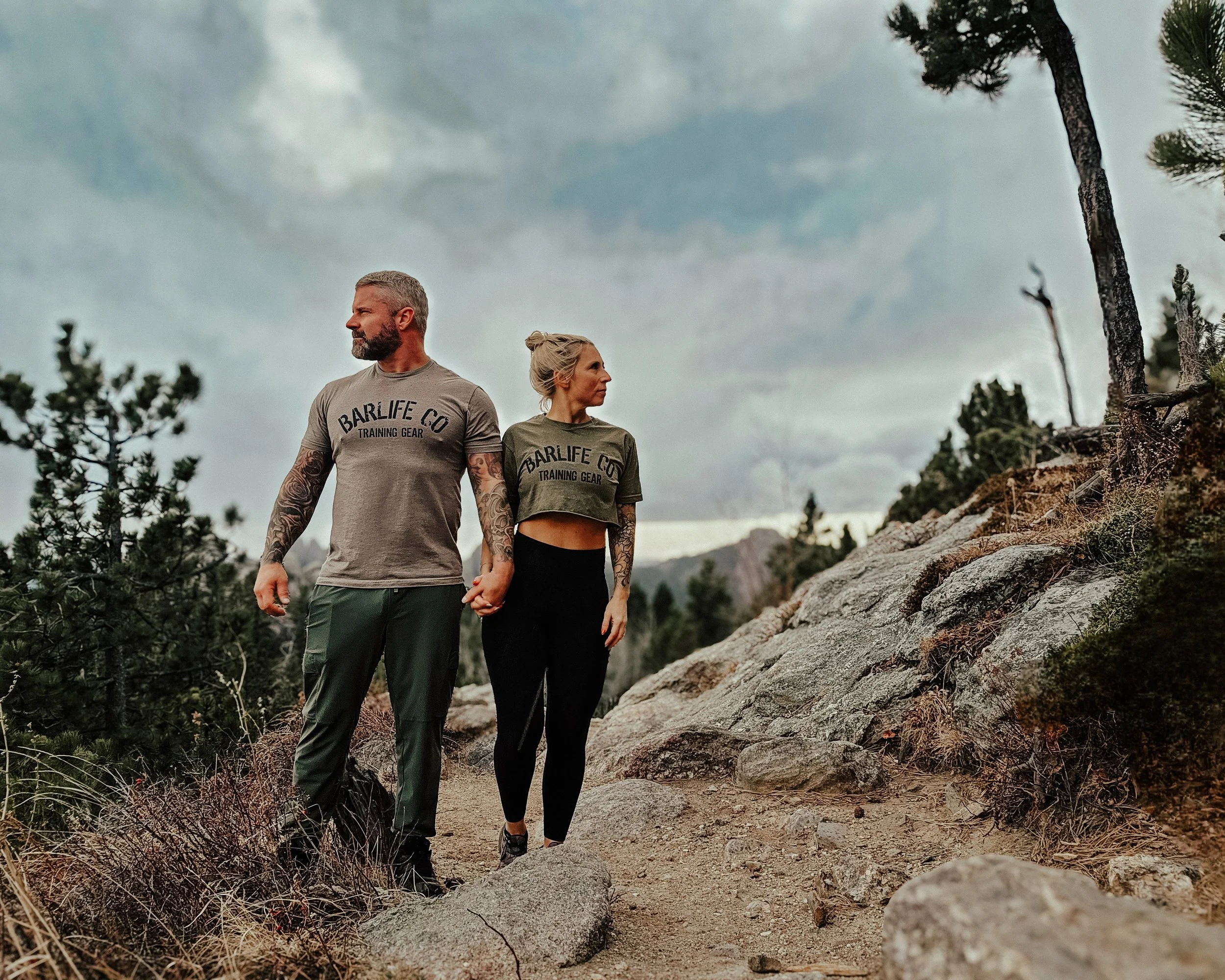 A couple holding hands and hiking on a rocky trail in a mountainous area with trees and cloudy sky.