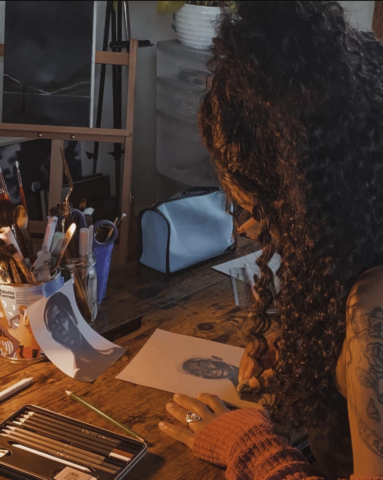 A woman with long, curly hair sitting at a wooden desk, drawing a portrait of a man with glasses on a piece of paper.