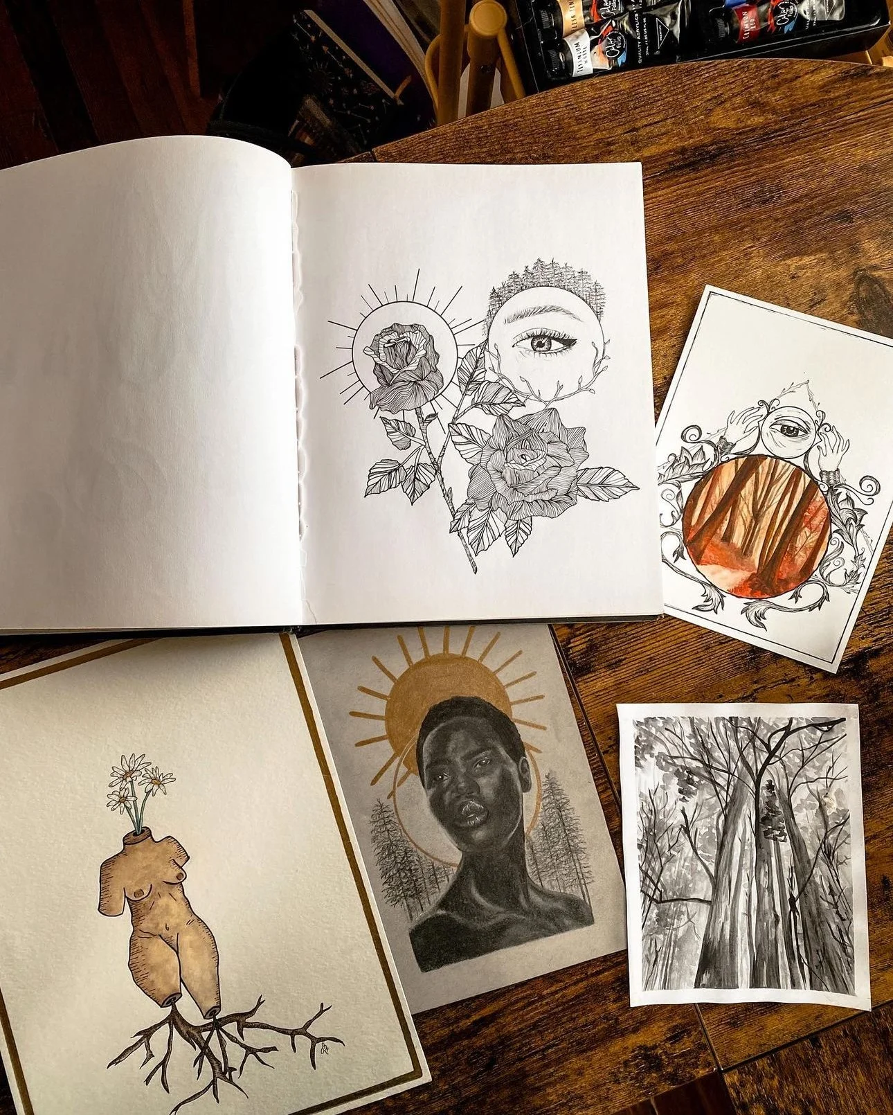 Several art prints and sketches on a wooden table, including a woman with a halo, a forest with an eye, a portrait of a dark-skinned woman with a sun halo, a tree with swing, and a cartoonish figure with flowers growing from its torso.