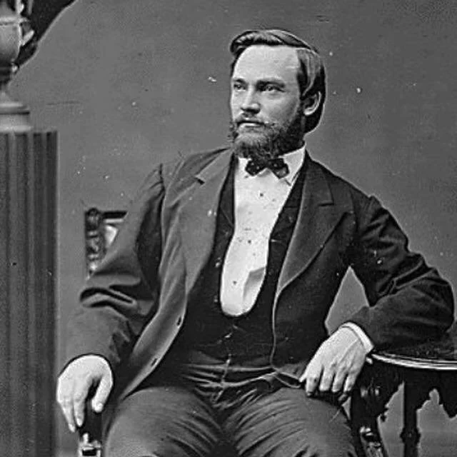 Black and white photograph of a man with a beard and mustache, sitting confidently, wearing a suit, bow tie, and white shirt, with one hand resting on a table and the other on his lap.