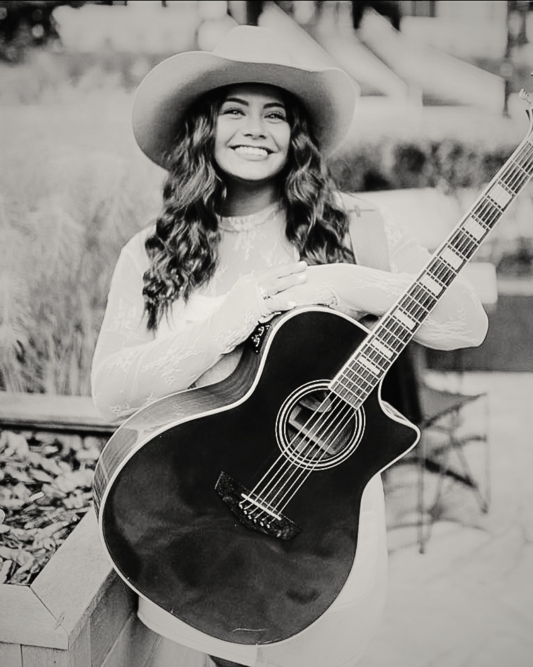 A woman with long, curly hair wearing a wide-brimmed hat and a lace top, smiling and holding an acoustic guitar outdoors.