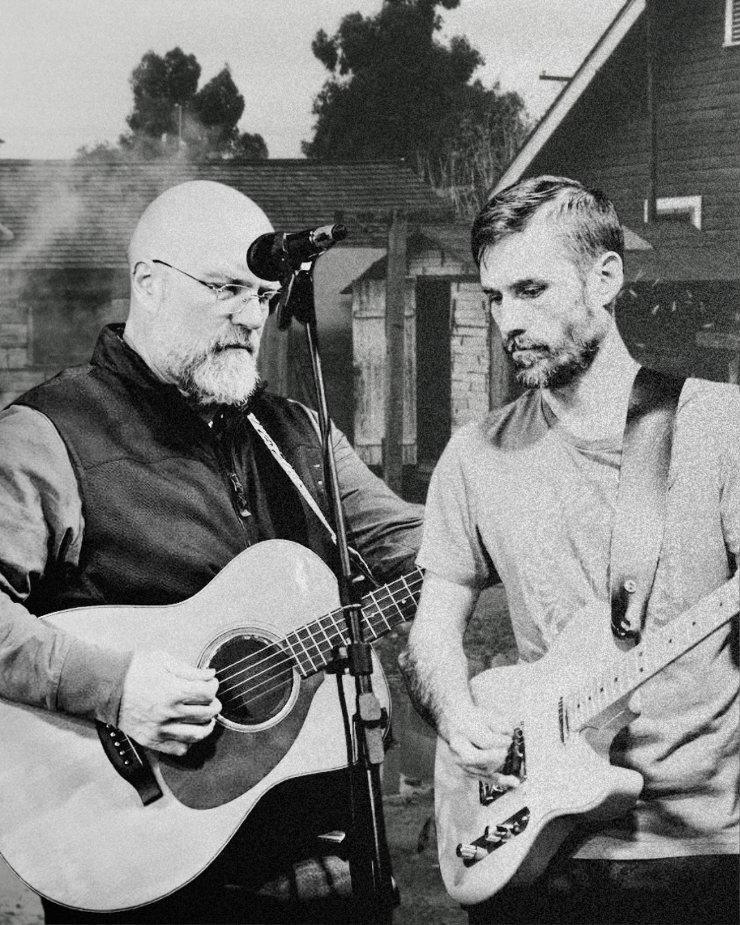 Two men playing guitars outdoors, one with a bald head and glasses, the other with short hair and a beard, focus on their instruments with a house and trees in the background.