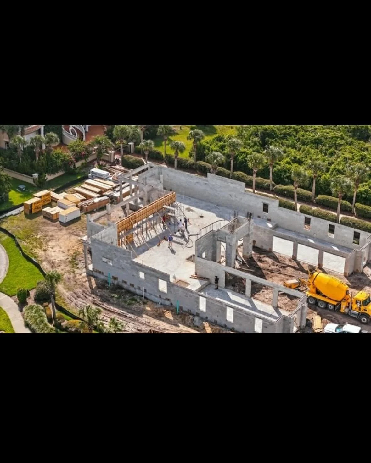 Aerial view of a building under construction with concrete walls, construction workers, a yellow cement mixer truck, and construction materials on site, surrounded by greenery.