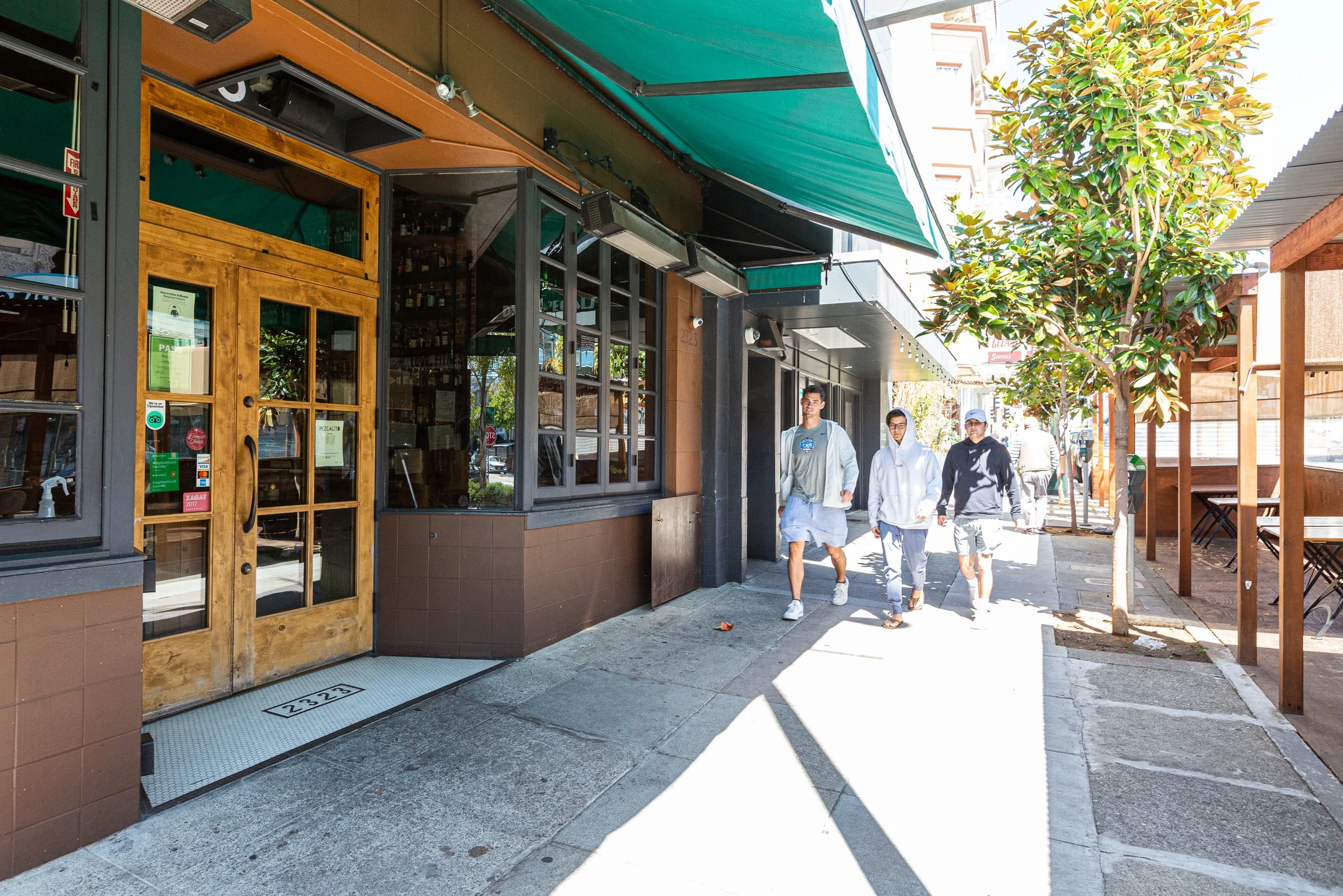 Pedestrians walking past a restaurant on a sunny city sidewalk with a green awning and a wooden door.