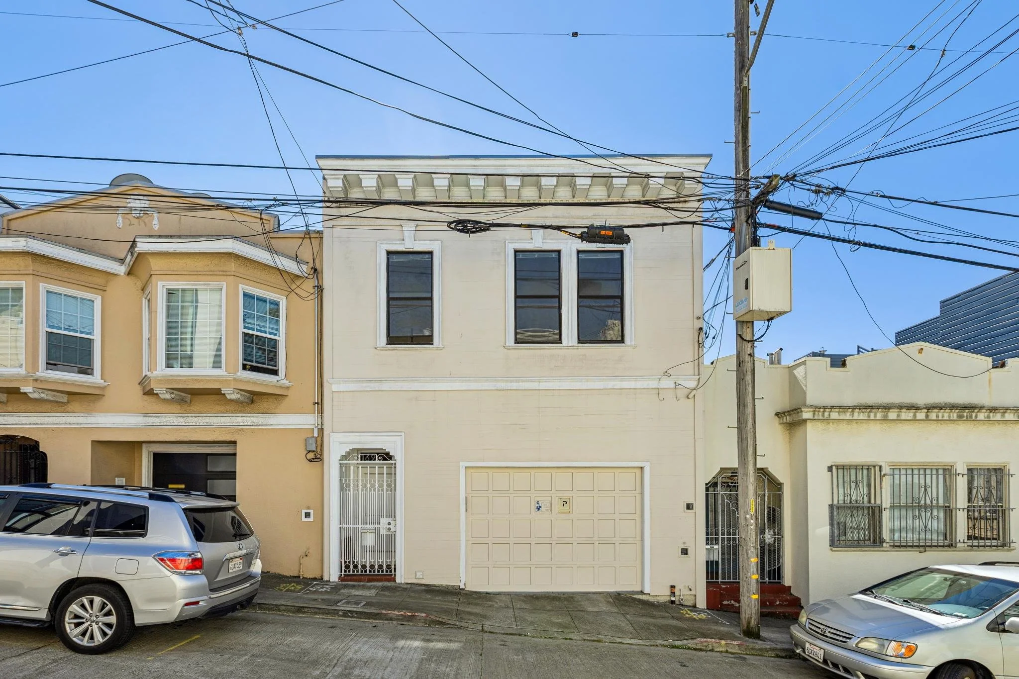 Street view of a two-story beige building with three black windows on the upper level, a white garage door, and a white front door, flanked by two neighboring buildings, with parked cars and a utility pole with numerous overhead wires.