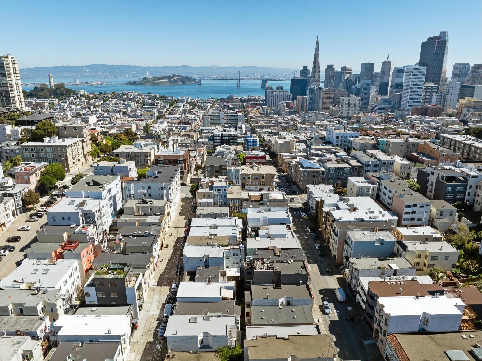 Aerial cityscape showing residential buildings, skyscrapers, a bridge, water, and hills in the distance on a clear day.