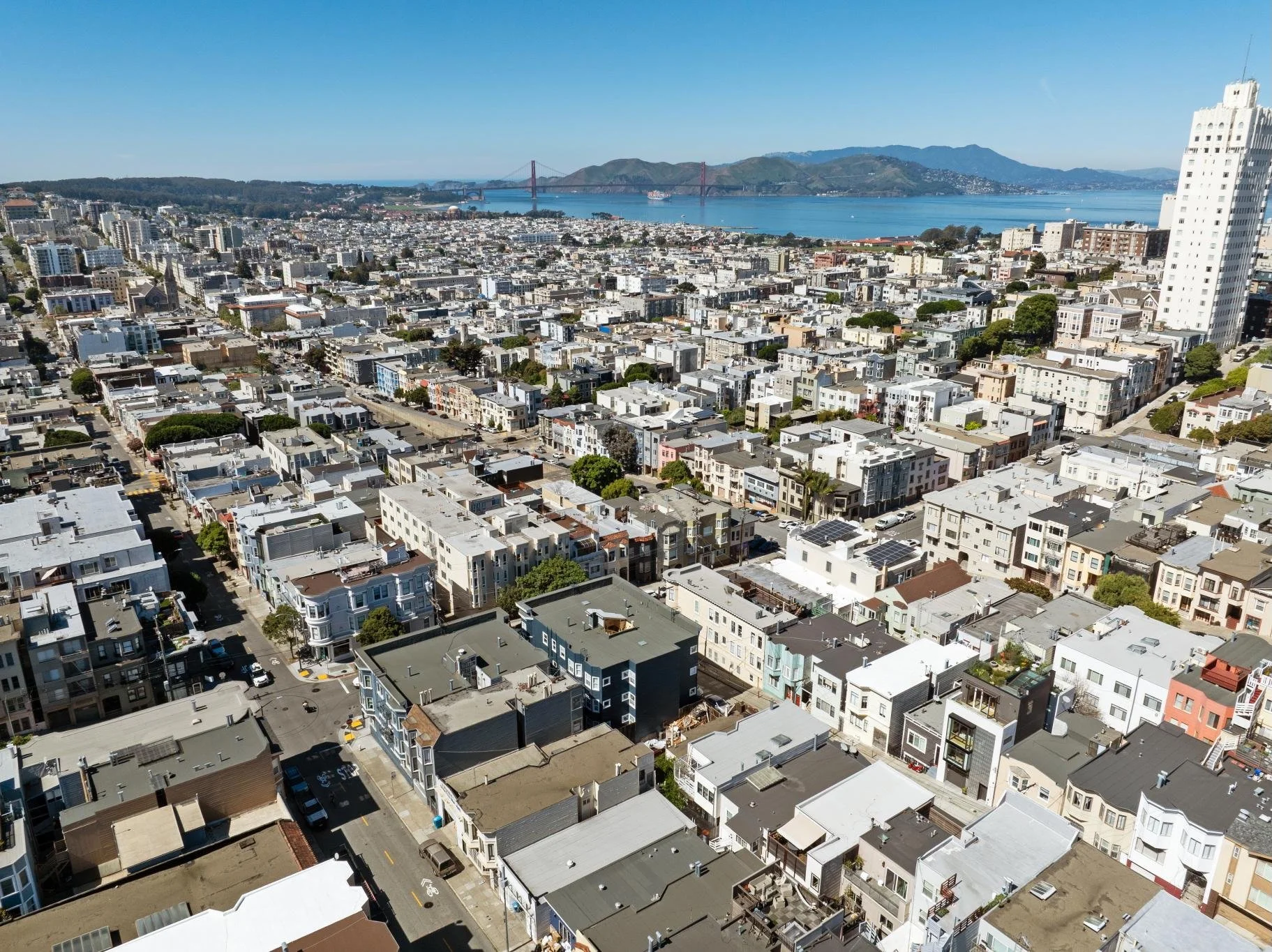 Aerial view of a cityscape with tightly packed buildings, streets, and green trees, with a large body of water and a bridge in the background under a clear blue sky.