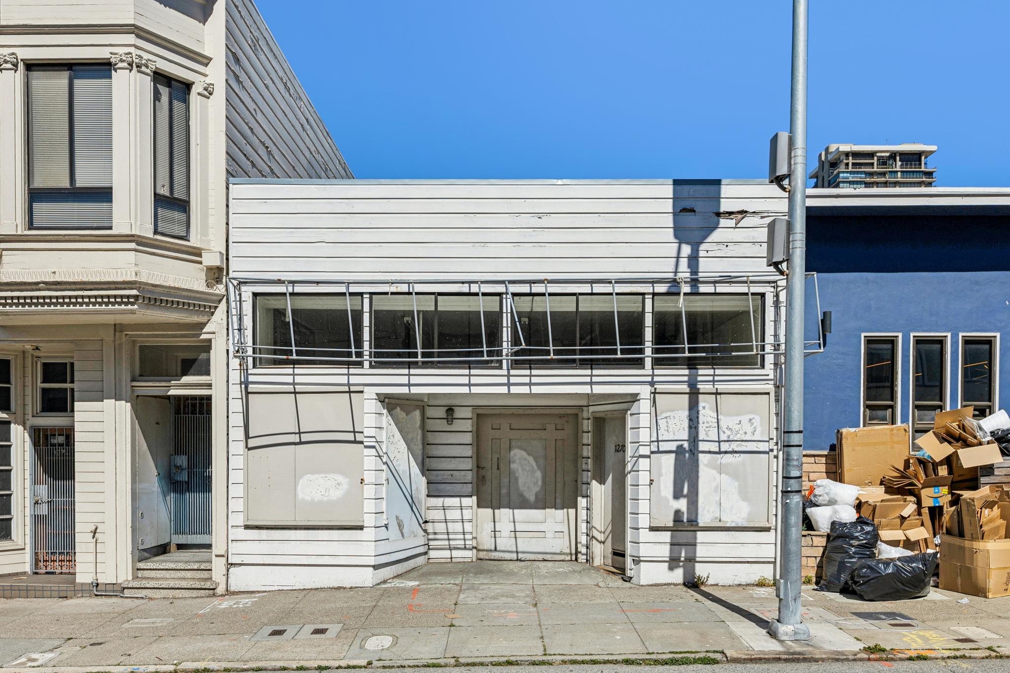 Empty white building with boarded-up windows and door, surrounded by trash bags and cardboard boxes on a city sidewalk, with a blue building to the right and a street lamp in front.