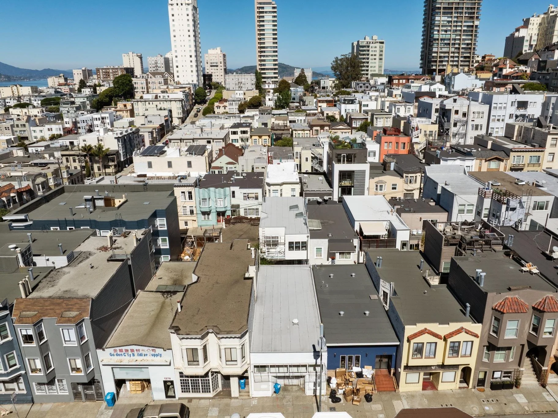 Aerial view of a densely packed urban neighborhood with multi-story residential buildings and shops, some with rooftop decks and solar panels, under a clear blue sky.