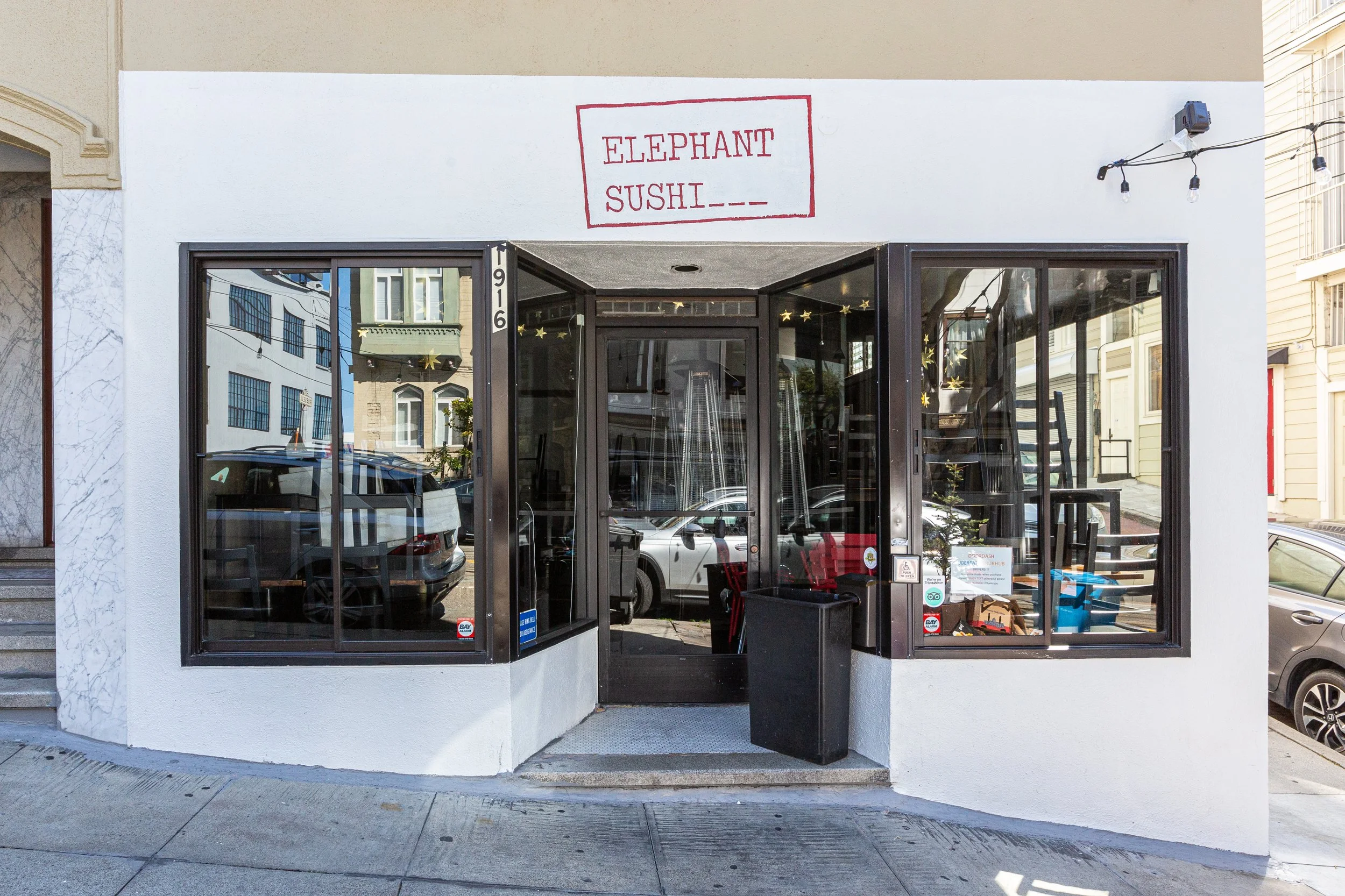 Front view of a small sushi restaurant named Elephant Sushi with a white exterior, black framed glass windows, and a welcoming entrance on a city street.