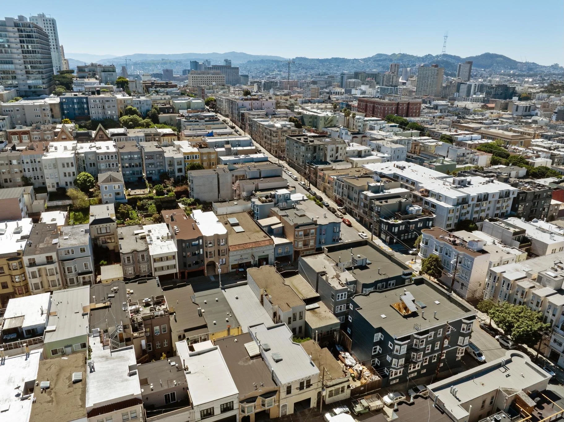 Aerial view of a city with tightly packed residential buildings, streets, and a few trees, with hills in the background under a clear blue sky.