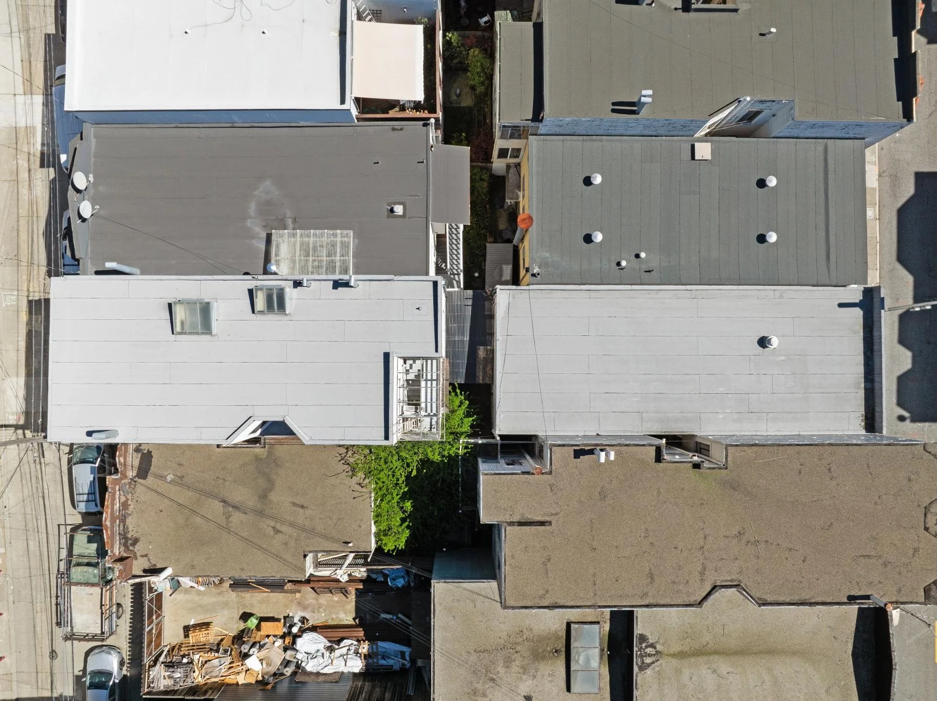 Aerial view of several rooftops in an urban area with different colors and shapes, some with vents and HVAC units, one with rooftop deck and greenery, and a small alley with construction materials and debris at the bottom.