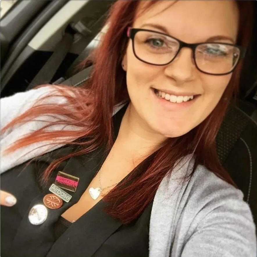 A woman with red hair, glasses, and a black attire, smiling for a selfie inside a vehicle.