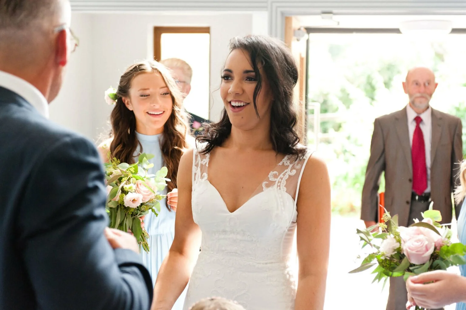 A bride with dark wavy hair in a white wedding dress smiling and holding hands with a man in a black suit in a wedding ceremony, surrounded by bridesmaids and guests inside a bright room with open doors.