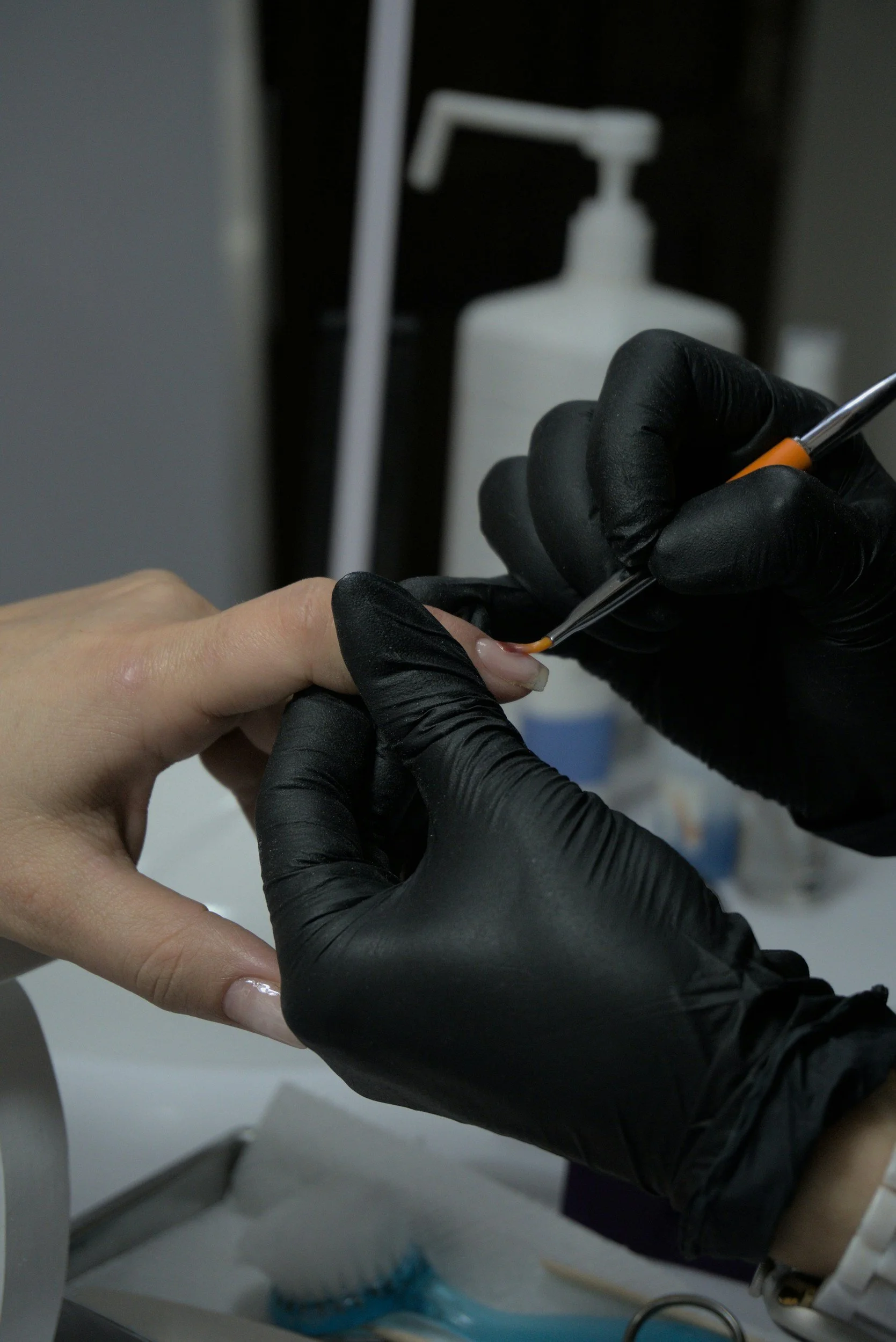 Close-up of a person with a light skin tone receiving a manicure, with a professional wearing black gloves using a tool on their fingernail. In the background, there is a white pump bottle, and the setting appears to be a salon or nail spa.
